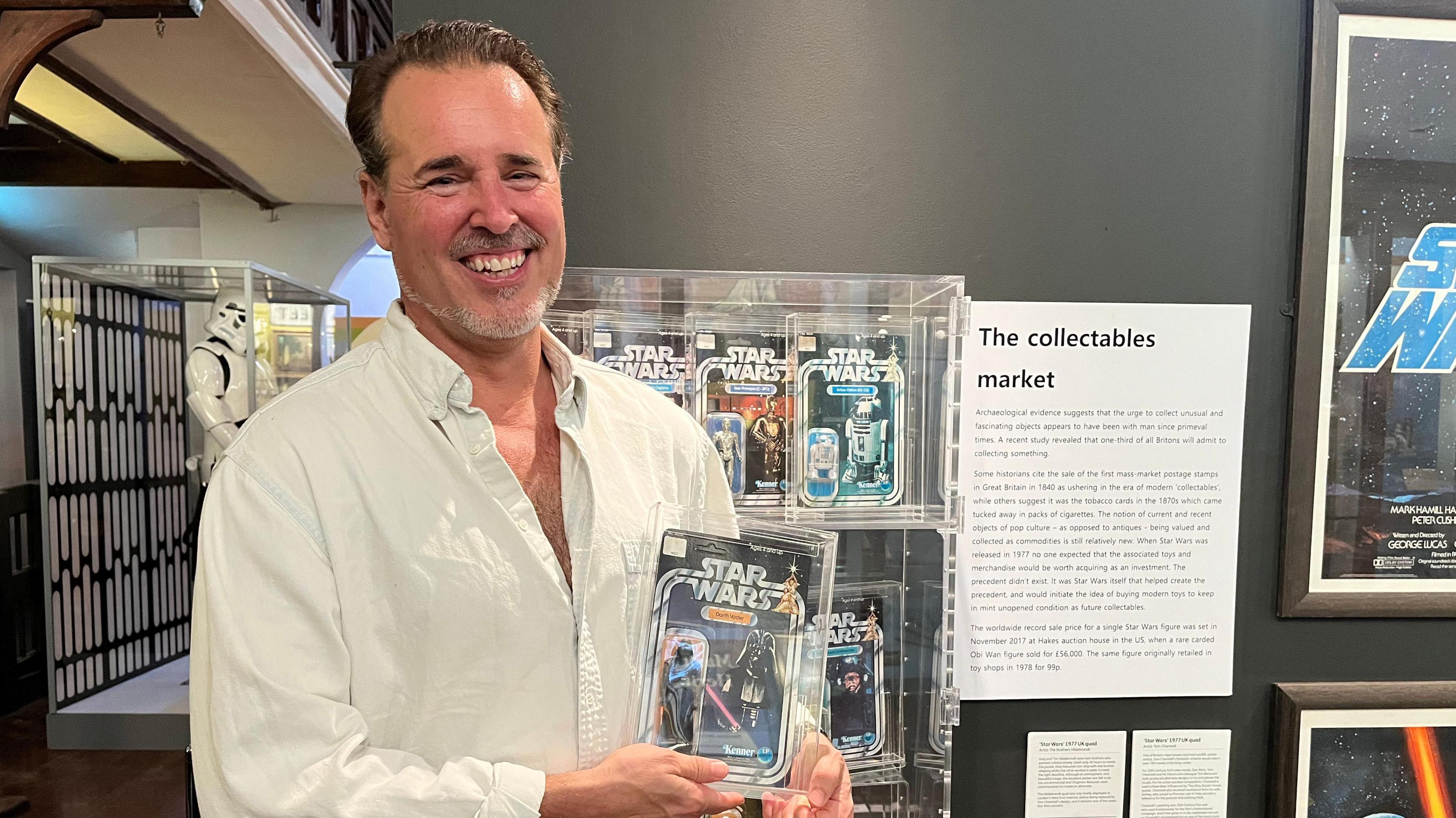 Matt Fox wears a white shirt and is standing in front of a wall of boxed star wars figurines. He is holding a Darth Vader box.