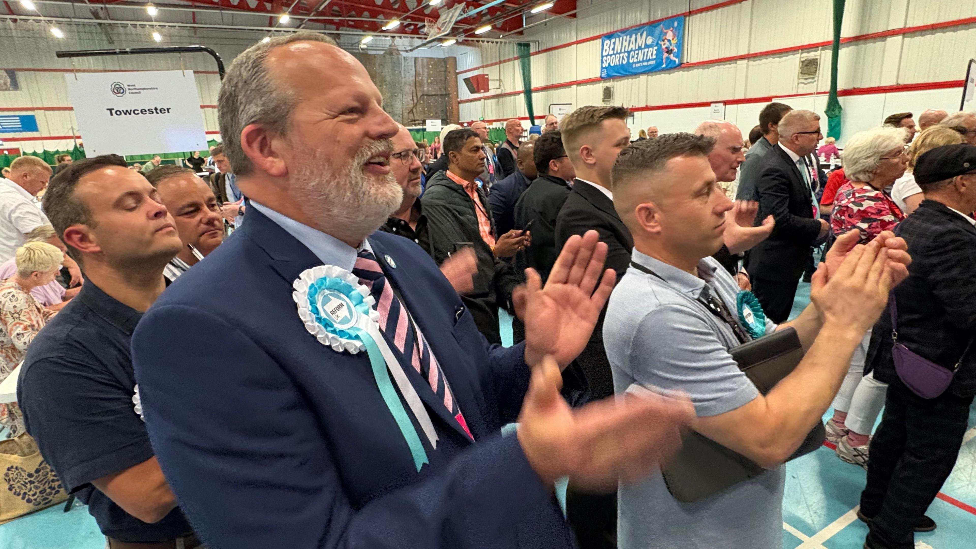 James Petter with short dark hair and grey beard, wearing a blue jacket, light blue shirt and pink and black tie, with a light blue reform rosette. He is smiling and clapping alongside other people similarly occupied at an election count.