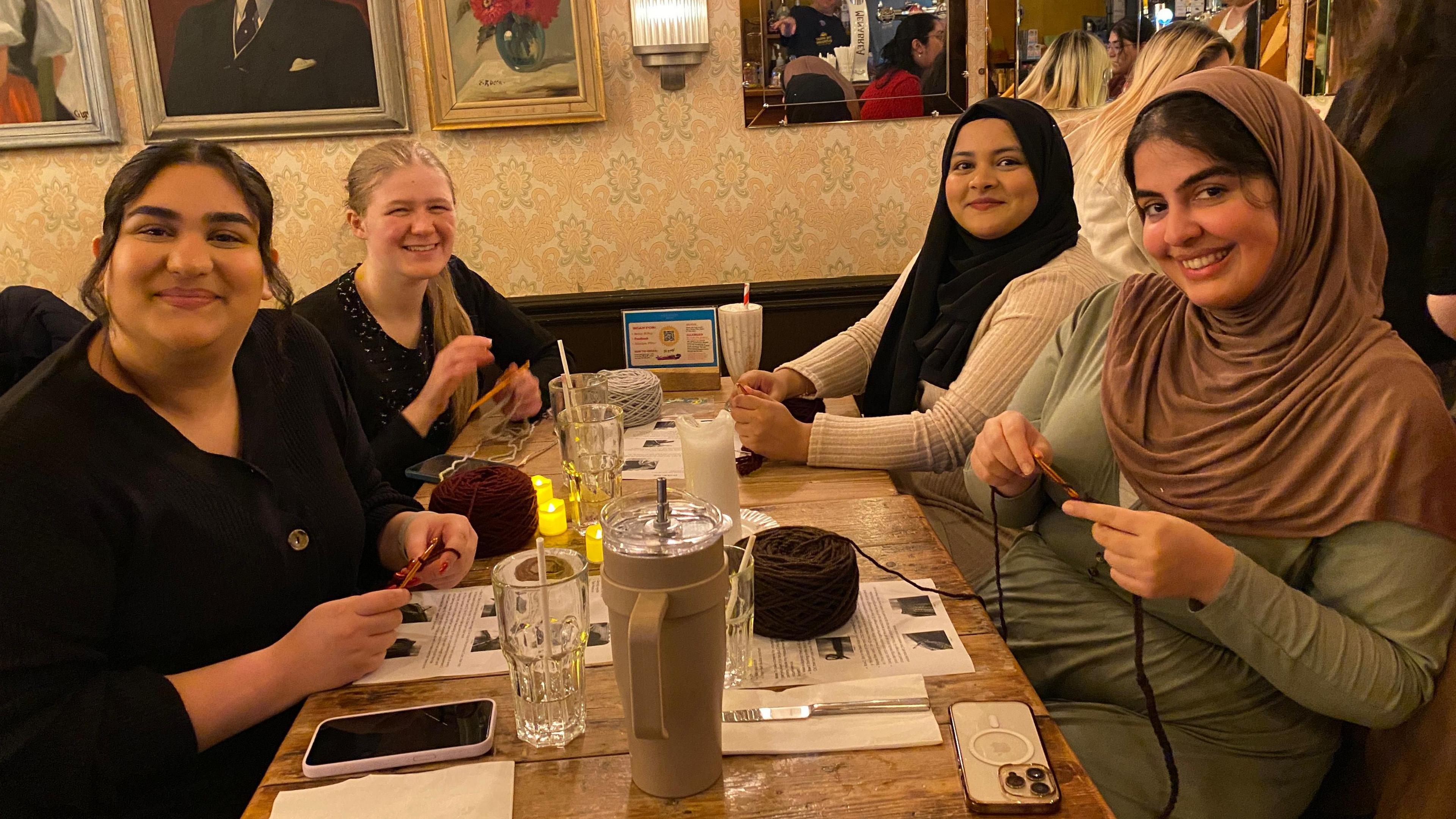 A group of four women are sitting around a table in a restaurant. They all have drinks on the table along with their phones as they learn to crochet with yarn