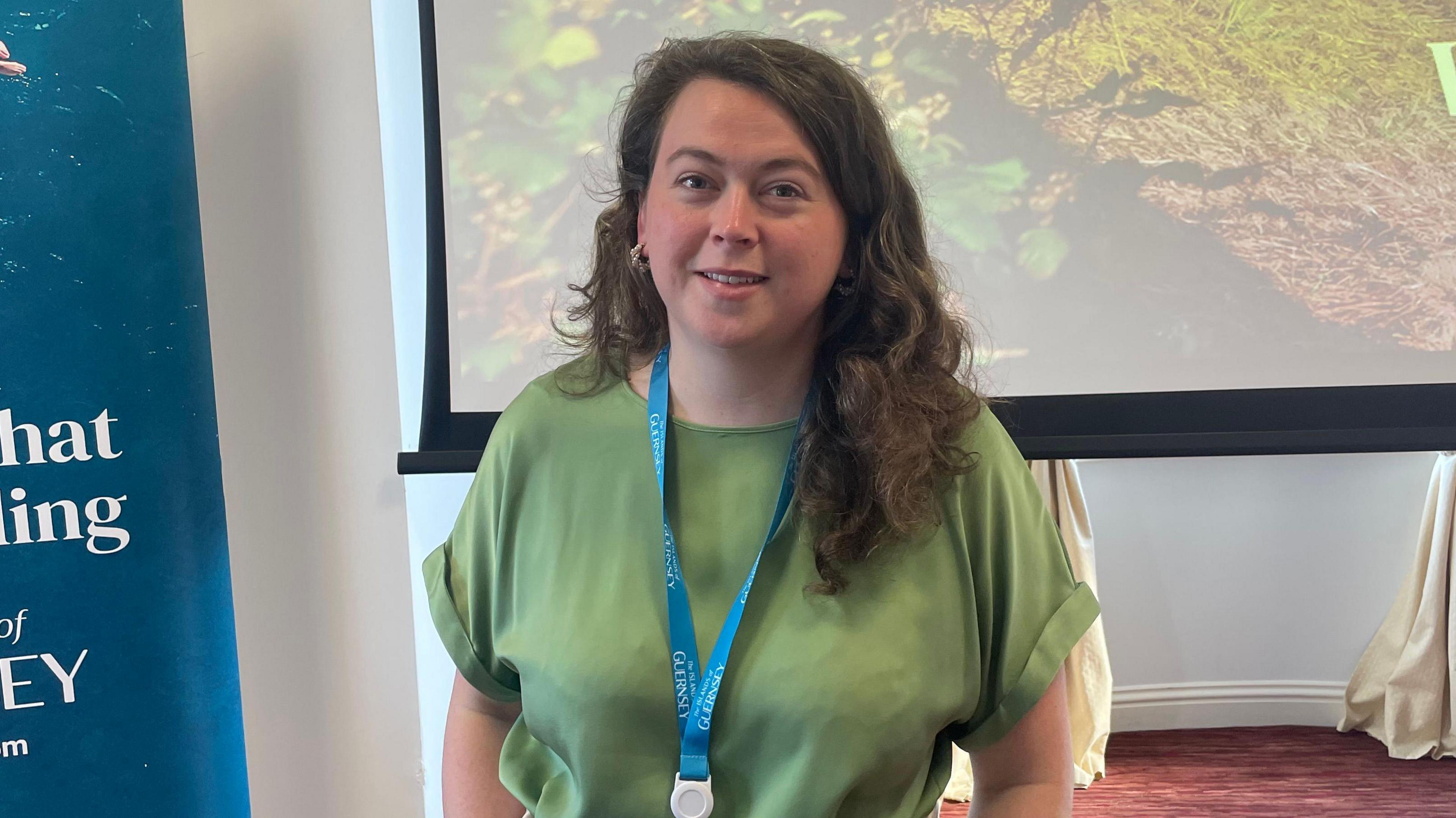 Jo Ferguson is wearing a green blouse and a turquoise lanyard. She has long dark hair and is smiling at the camera. She is standing in front of a giant screen which has a photos showing shrubs and grass. To her right is part of a banner.