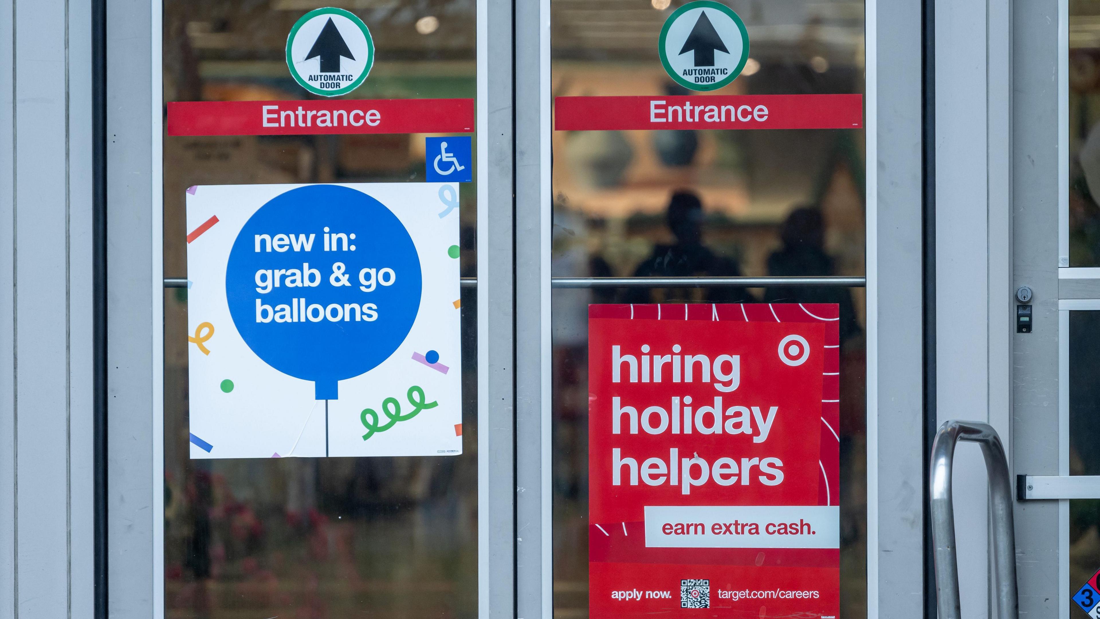 A red sign reading "Hiring Holiday Helpers" hangs on a glass entry door to a Target store.