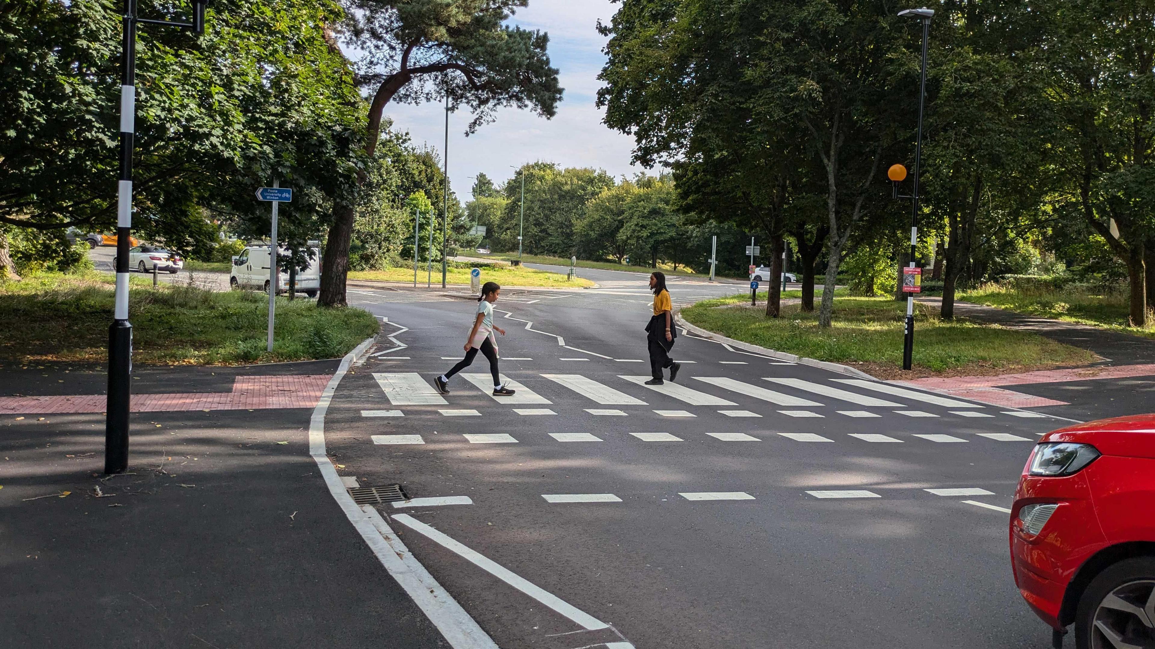 The new crossing on Glenferness Avenue looking towards Talbot roundabout. Two people are seen crossing the road. Part of a red car is seen waiting. It's a sunny day.