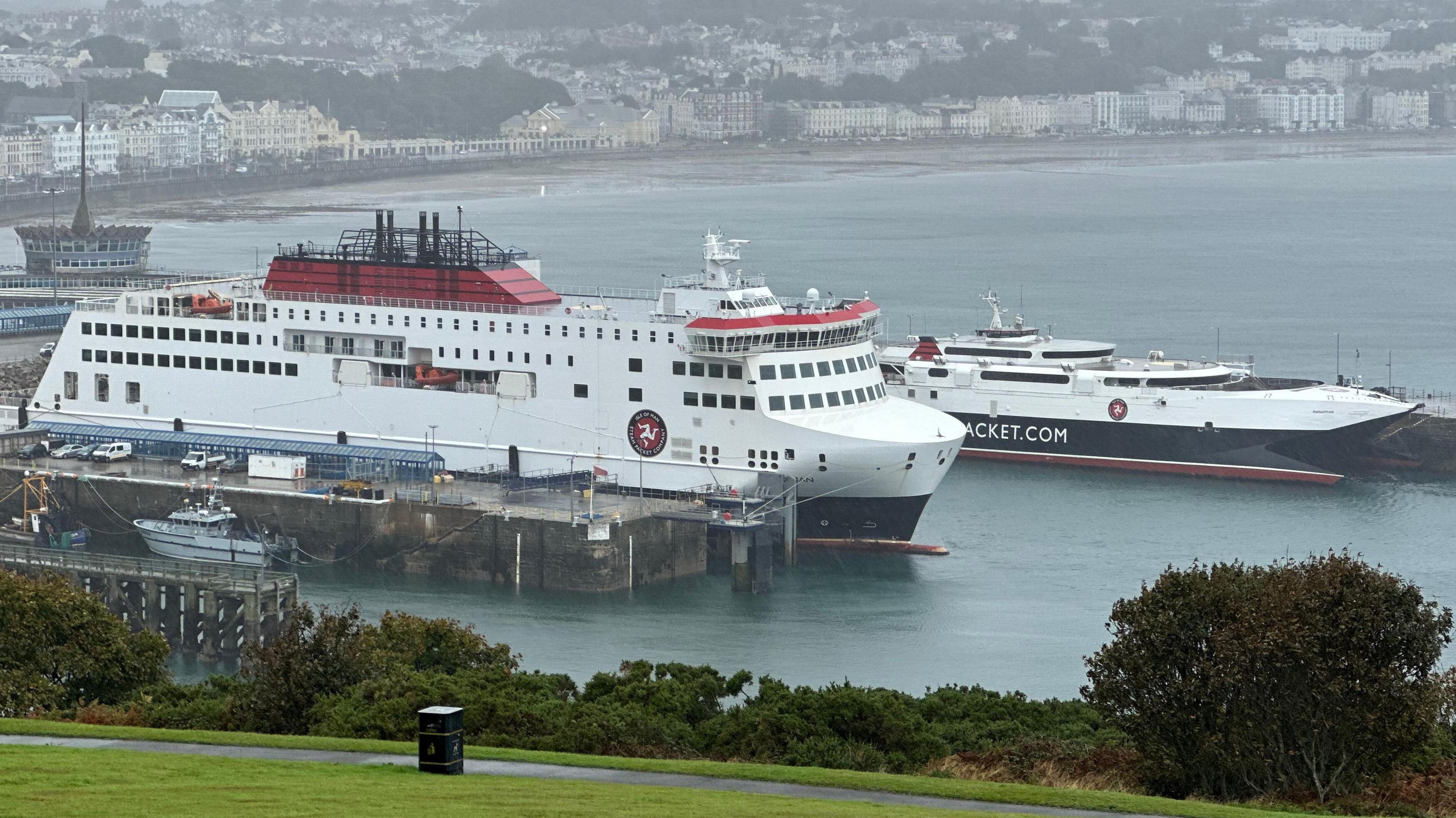 The Manxman and Manannan ferries moored in Douglas Harbour. The are painted in the colours of the Steam Packet Company of red, white and black. The sweep of Douglas Bay the buildings on Douglas Promenade provide a backdrop to the vessels.
