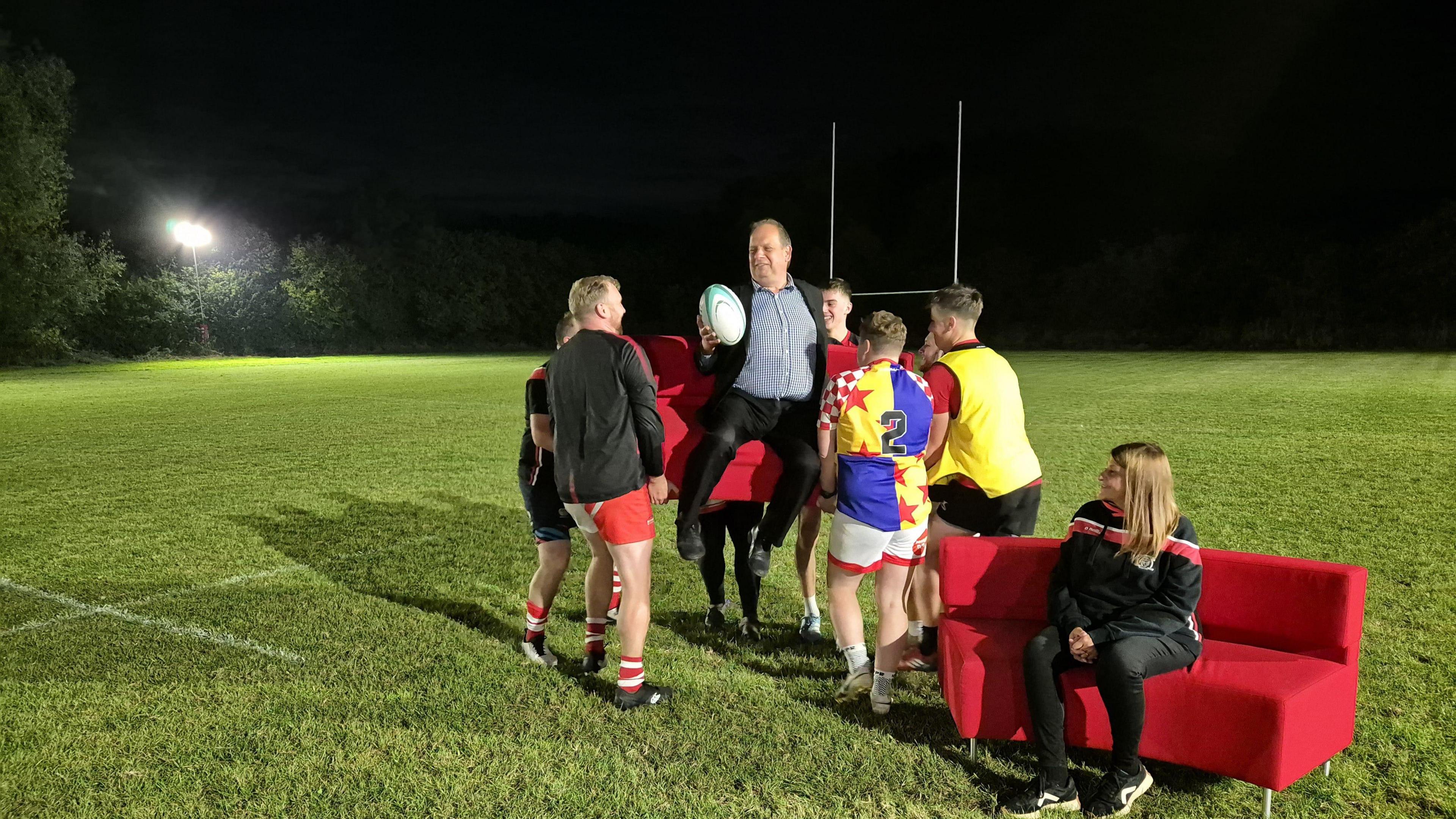 Rugby players at Claverdon RFC lift the red sofa, with reporter Ben Sidwell sitting on it, and carry it across the pitch