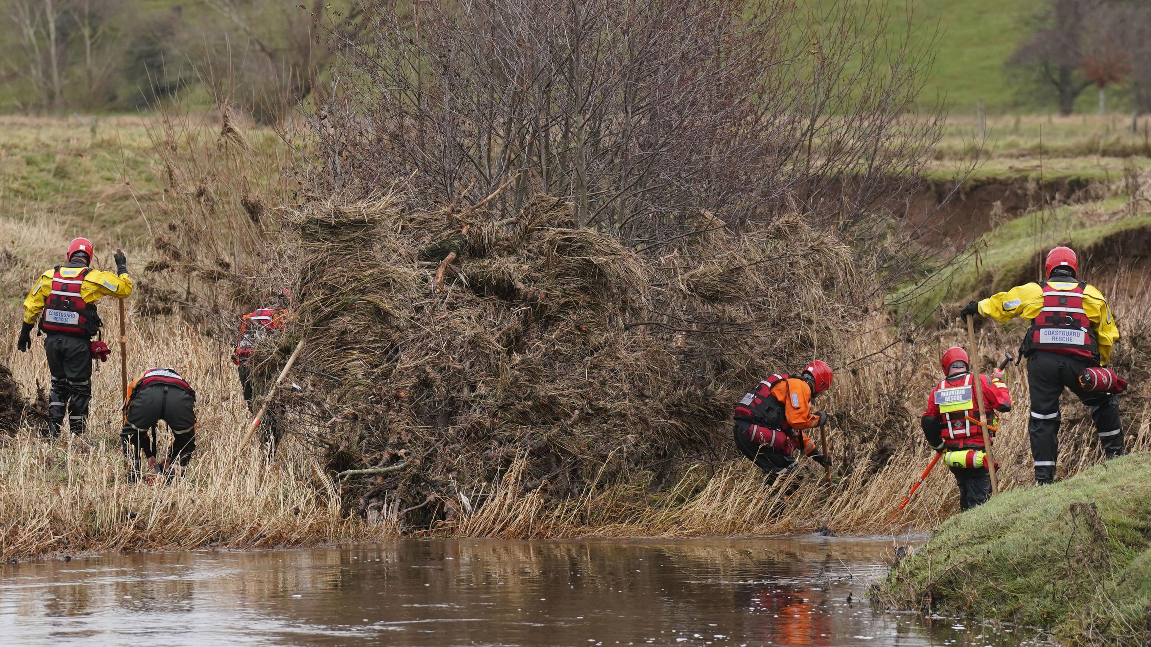 Several people wearing waterproof gear and lifejackets and helmets are scouring the river bank. There is debris on the bank.