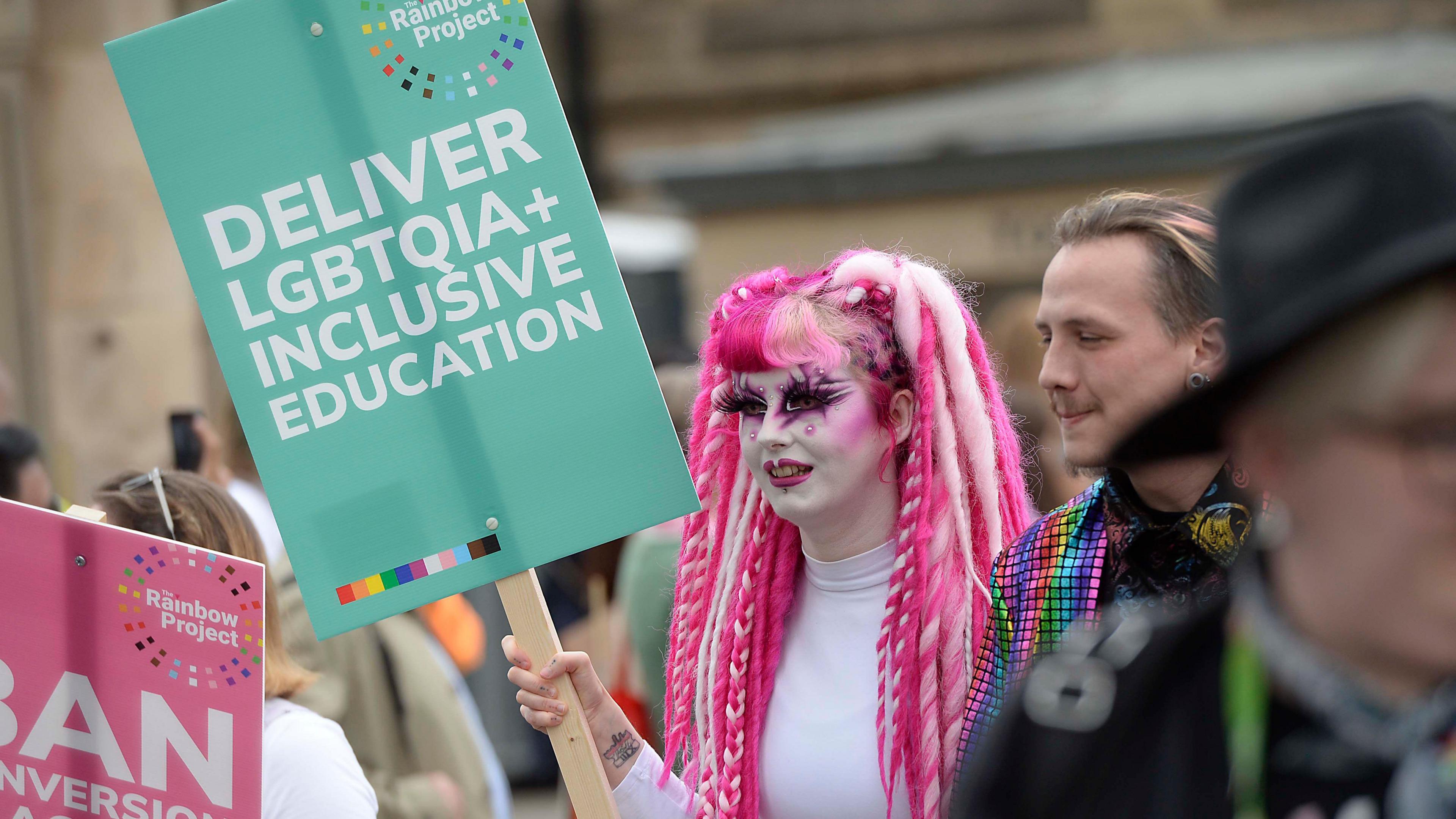 Person carrying a sign that reads "Deliver LGBTQIA+ Inclusive Education"