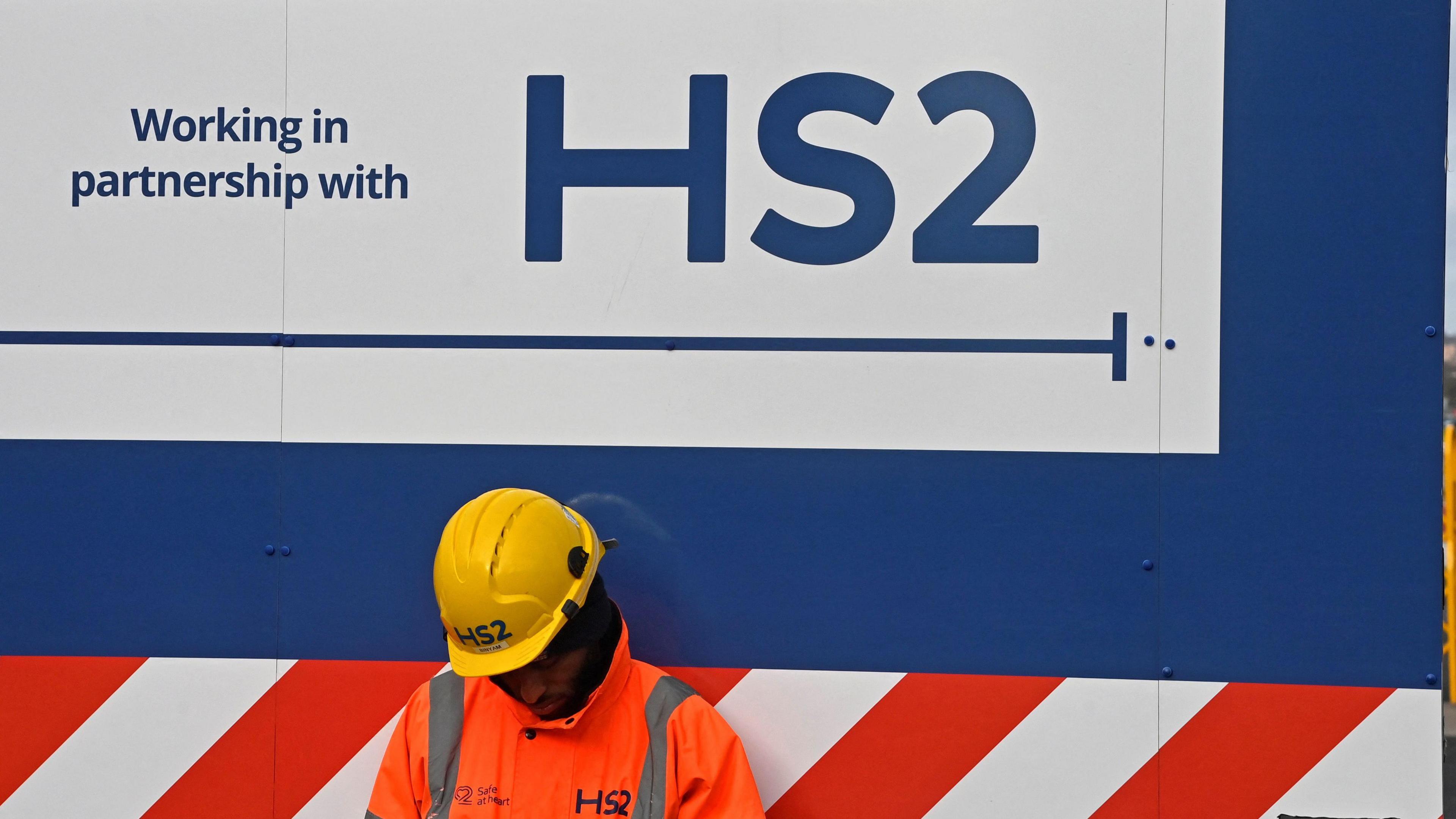 An HS2 workers in orange hi-vis clothes and a yellow hard-hat sits in front of blue, red and white hoarding with an HS2 sign on it