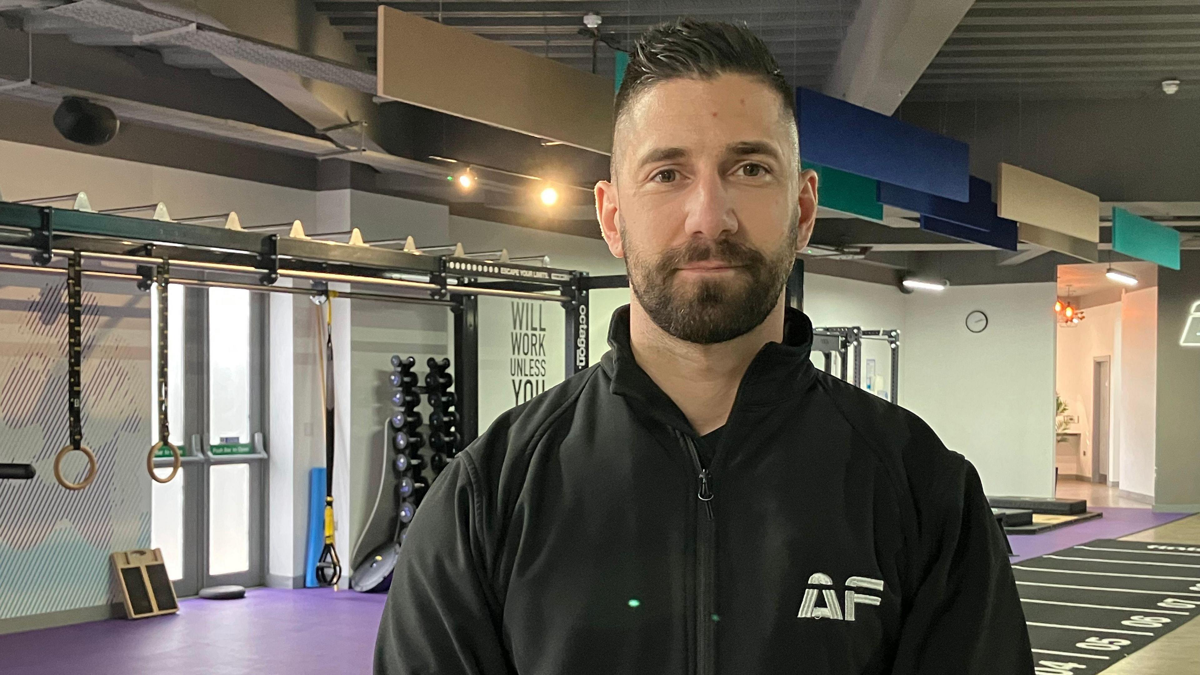A man wearing a black zip up hoodie. Behind him is gym equipment. The lights are on. He is looking at the camera and has black hair and a black beard.