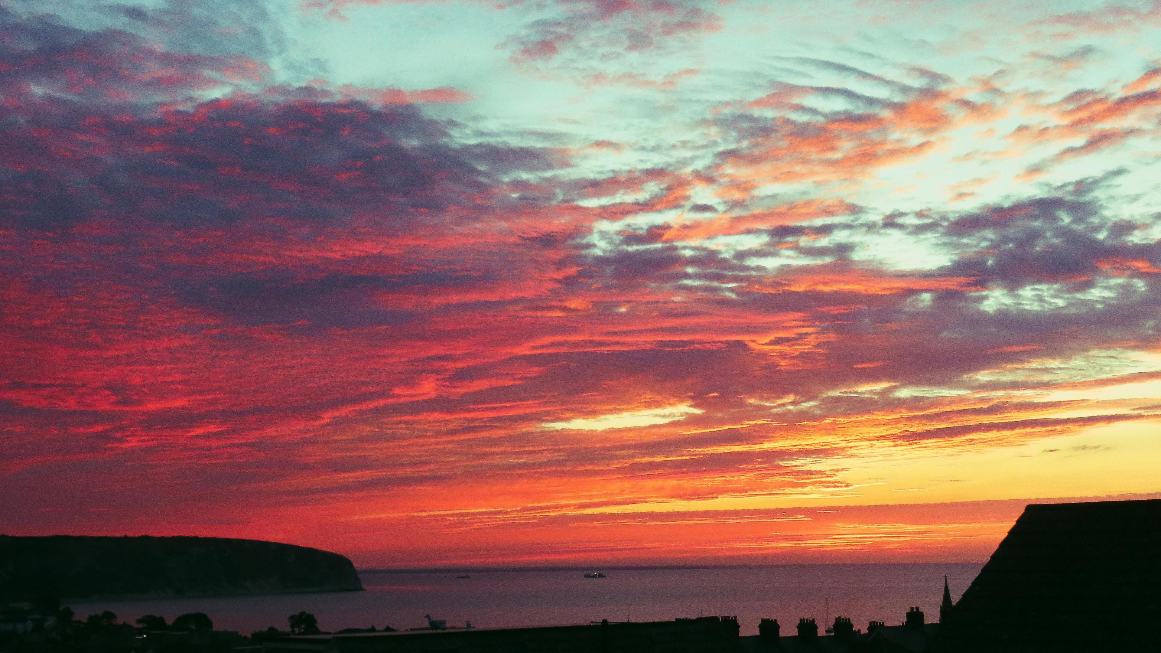 Sunrise over the coast with the sky bursting with orange and yellow hues through cloudy skies moving away revealing blue skies. A house and coastline can be seen in silhouette.