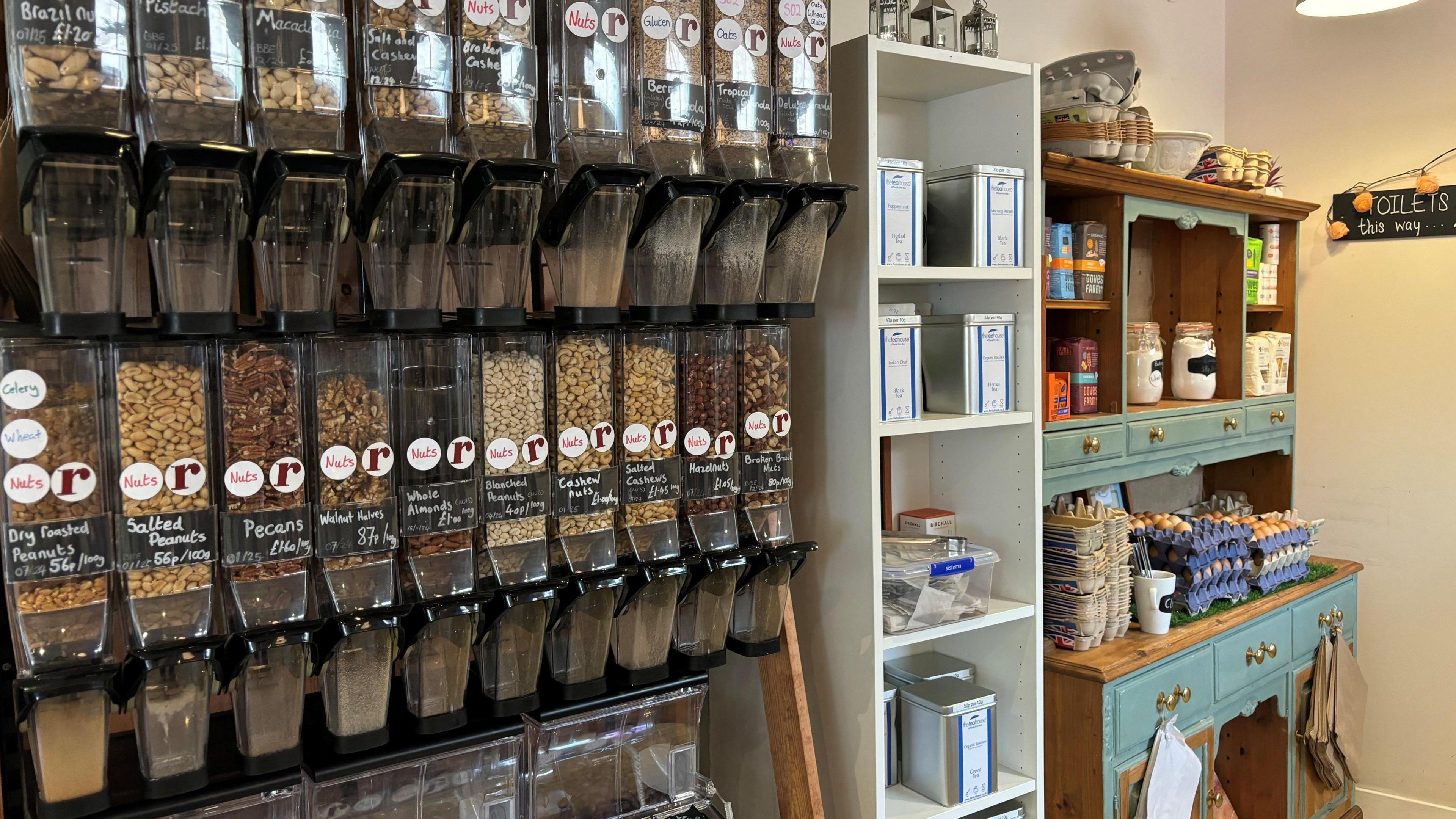 The inside of Roots cafe. The wall is covered with refill containers of nuts, beans, seeds and cereals. The is a white shelving unit in the middle with metal tins of flour and other dry ingredients, and on the right there is a blue dresser with stacks of egg cartons. 