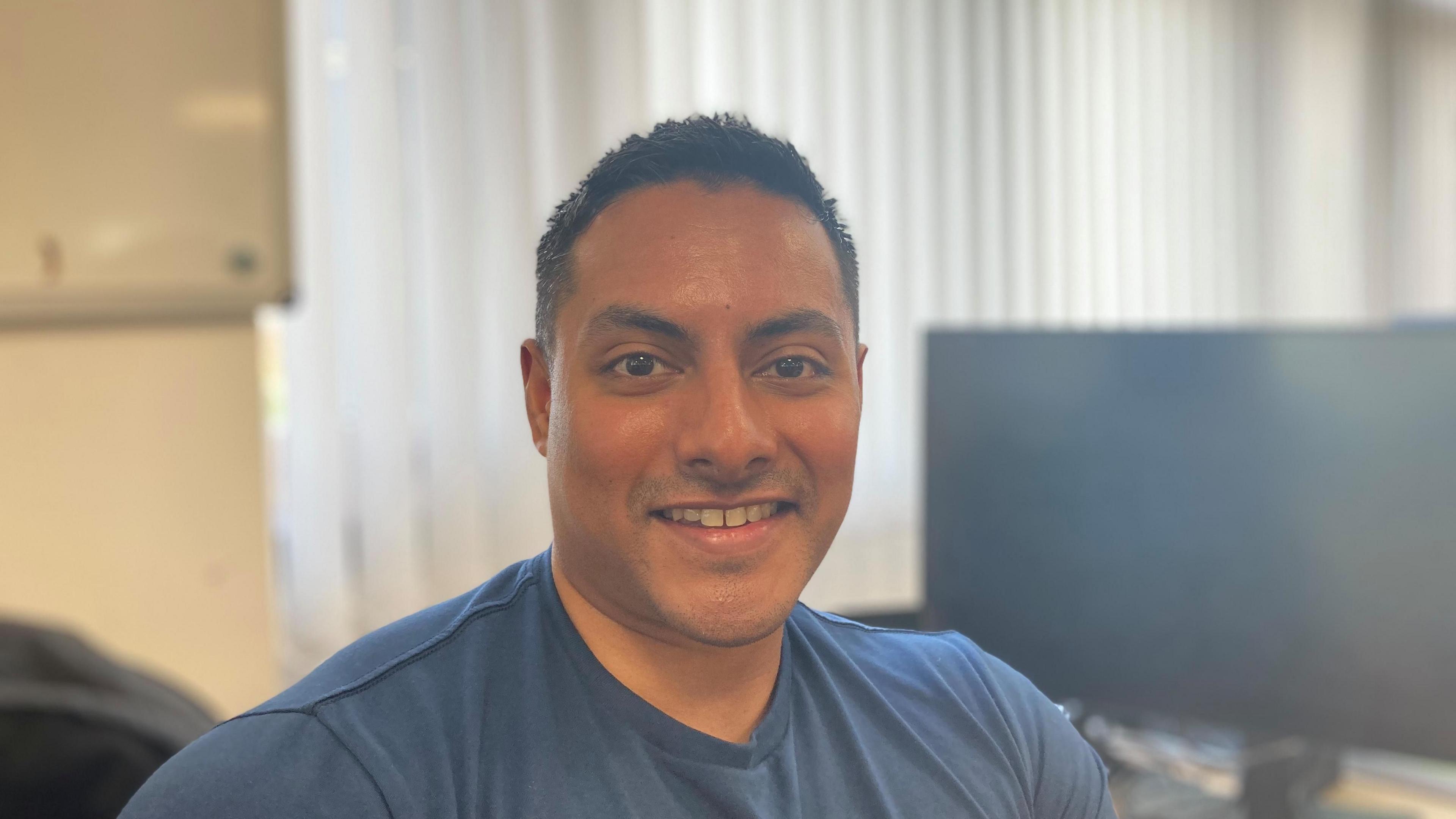 Police Sergeant Abdul Salique looking at the camera smiling. He is wearing a blue T-shirt and sat in front of a computer monitor