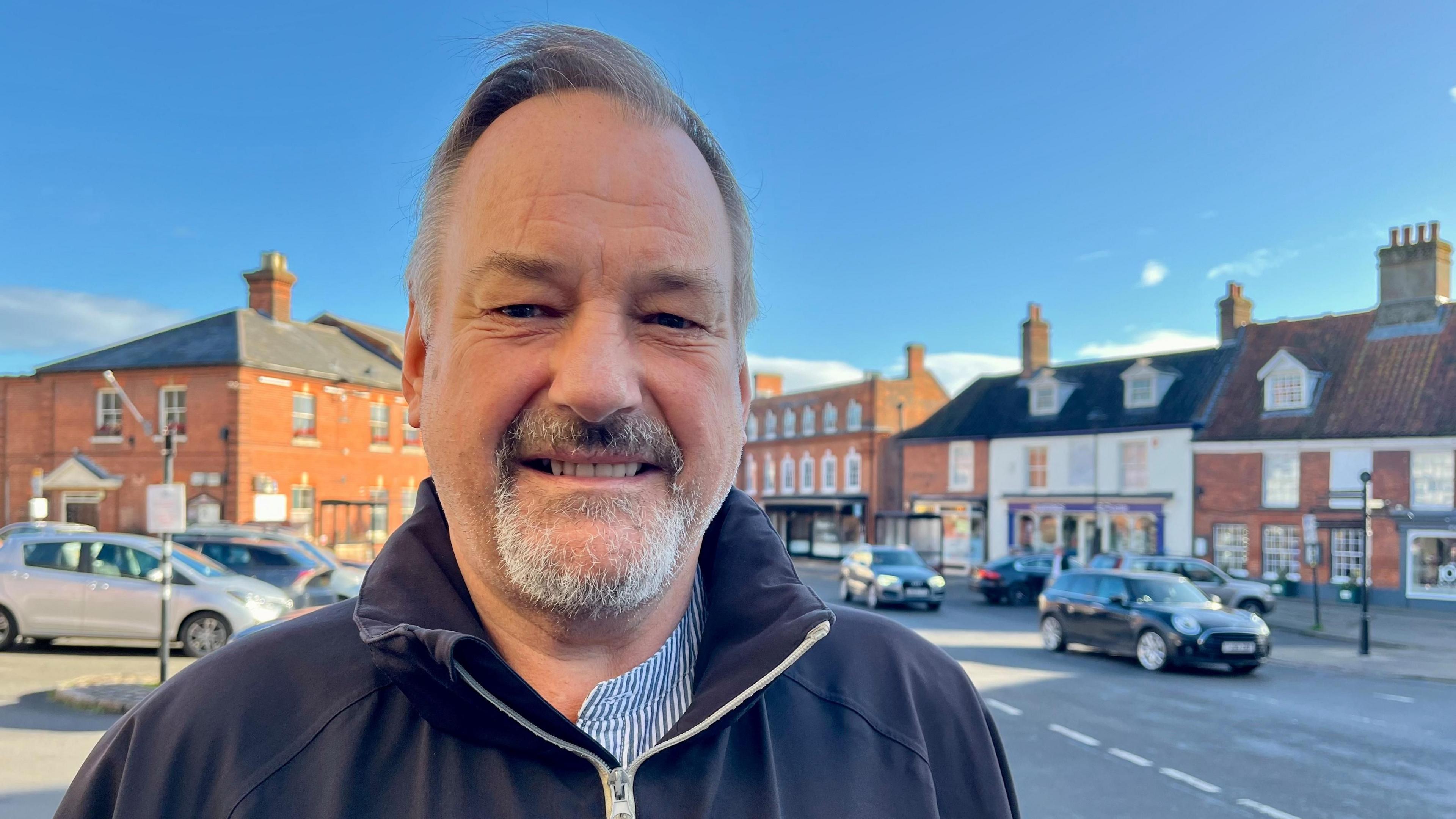 Martin Skoyles has short, grey hair and a small, grey beard. He is wearing a blue jumper with a striped blue shirt underneath.
He is standing in Aylsham market place with parked cars and shops behind him.