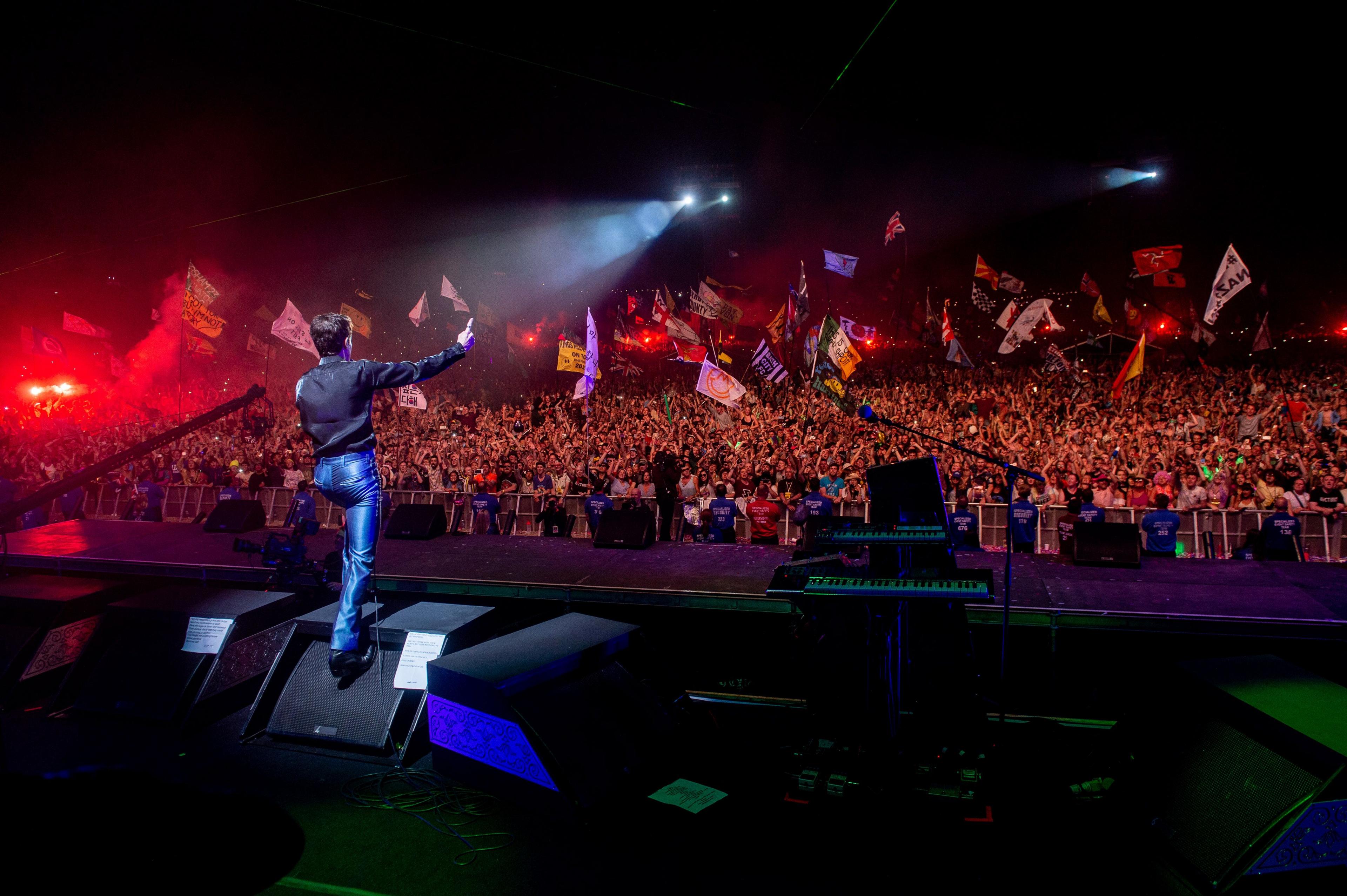 The Killers playing Glastonbury's Pyramid Stage. The photo is taken from the back of the stage, showing Brandon Flowers silhouetted against thousands of flag-waving fans