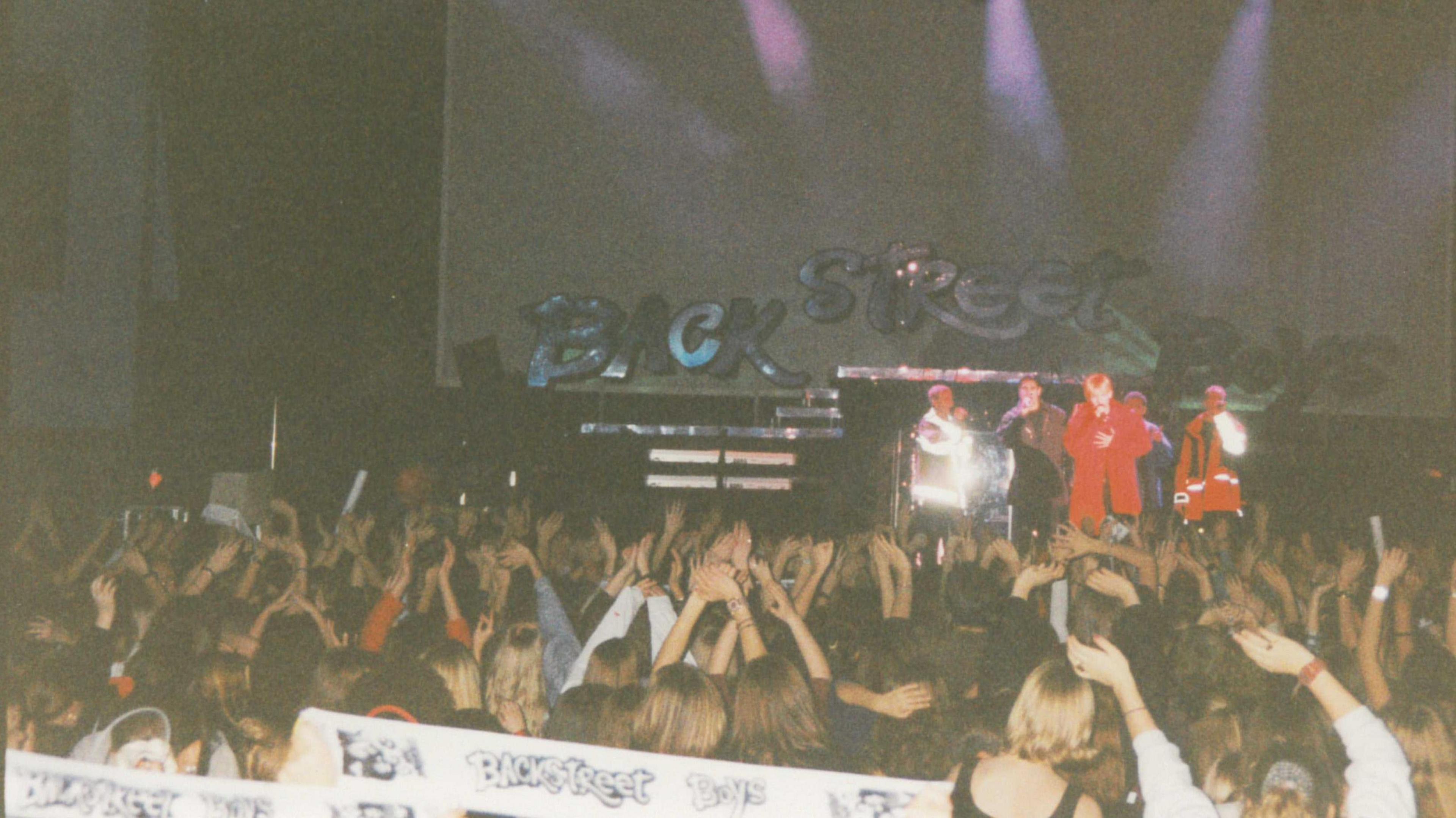 A grainy photograph showing the Backstreet Boys on stage with the band name behind them in silver letters. The photo is taken from far back in the crowd, which can be see with their hands in the air and holding up Backstreet Boys scarves