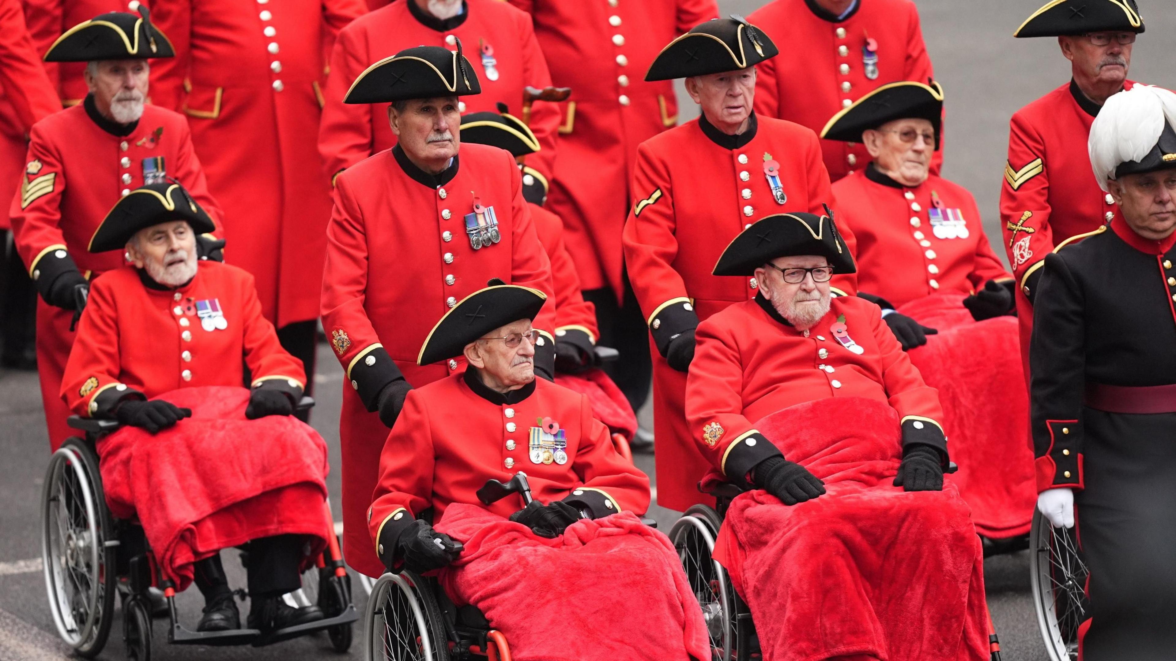 A group of eight Chelsea pensioners parade down the mall in central London for the Remembrance Day event. They are dressed in long red coats, medals, and tricorner hats. 