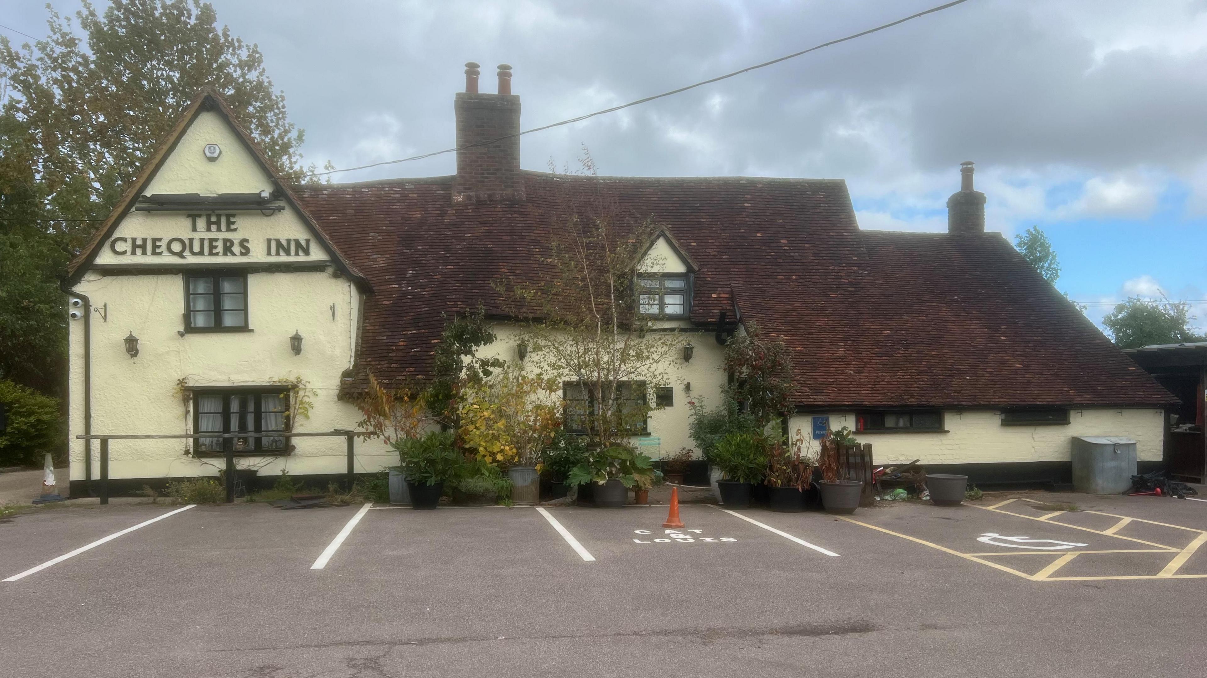 The front of the Chequers Inn before the blaze, with greenery outside and a tiled roof that has bowed over the centuries. There are empty parking spaces outside. One of the spaces is marked for disabled users and another space has a traffic cone on it.