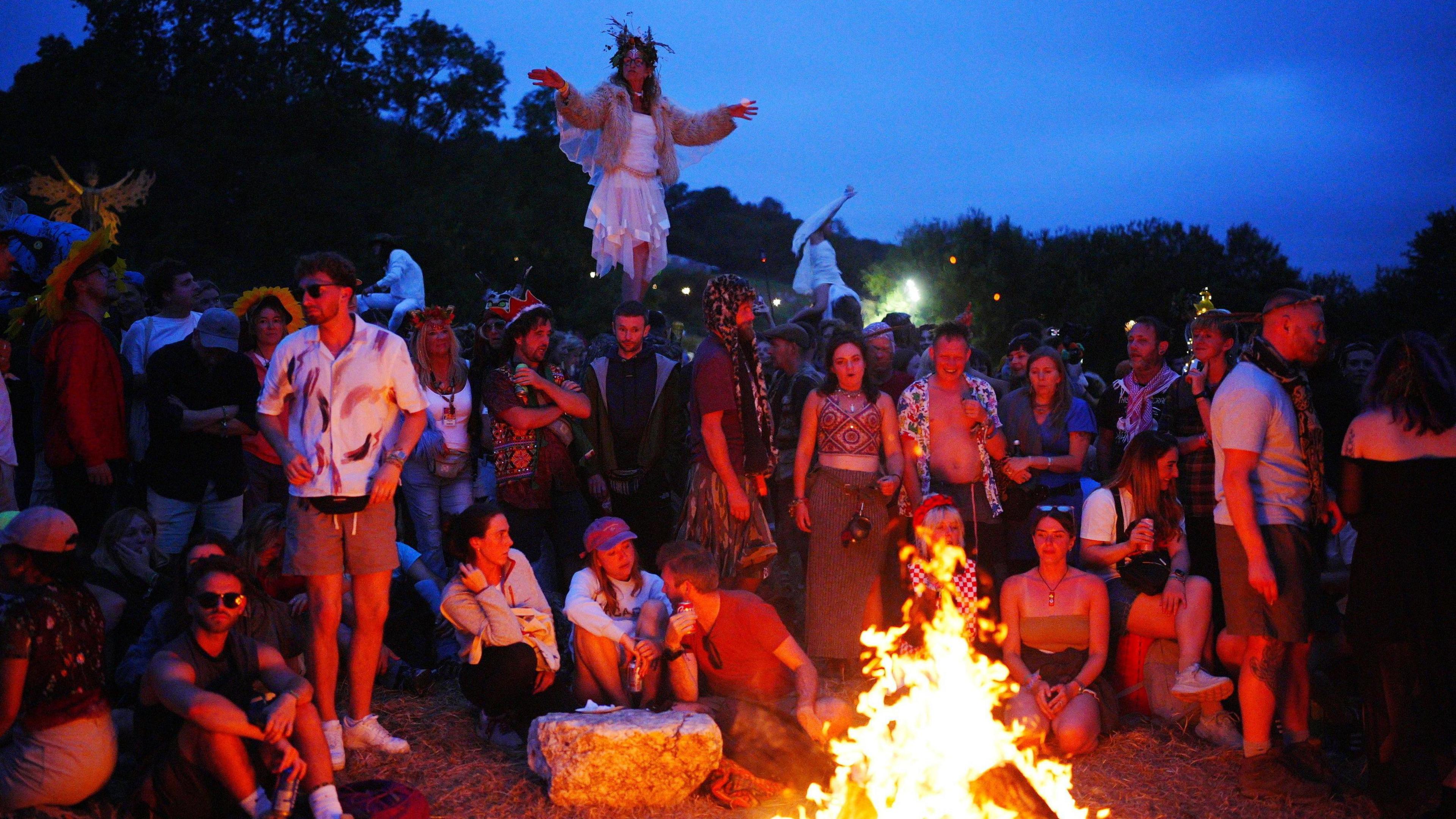 Revellers at the Stone Circle enjoying Glastonbury Festival. There is a fire in the foreground which people are gathered around.