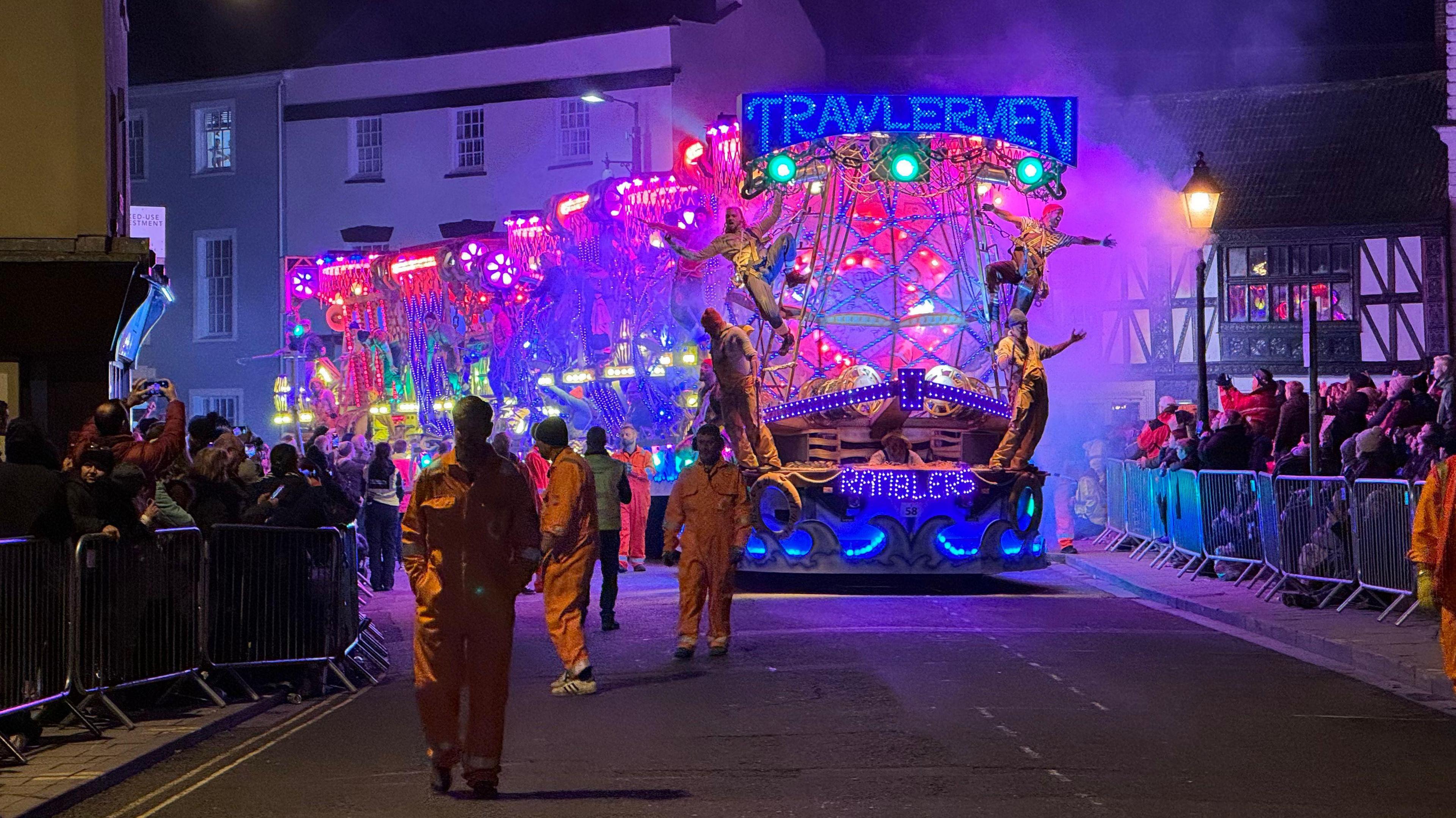 An illuminated cart with performers dangling off the sides proceeds through a high street.