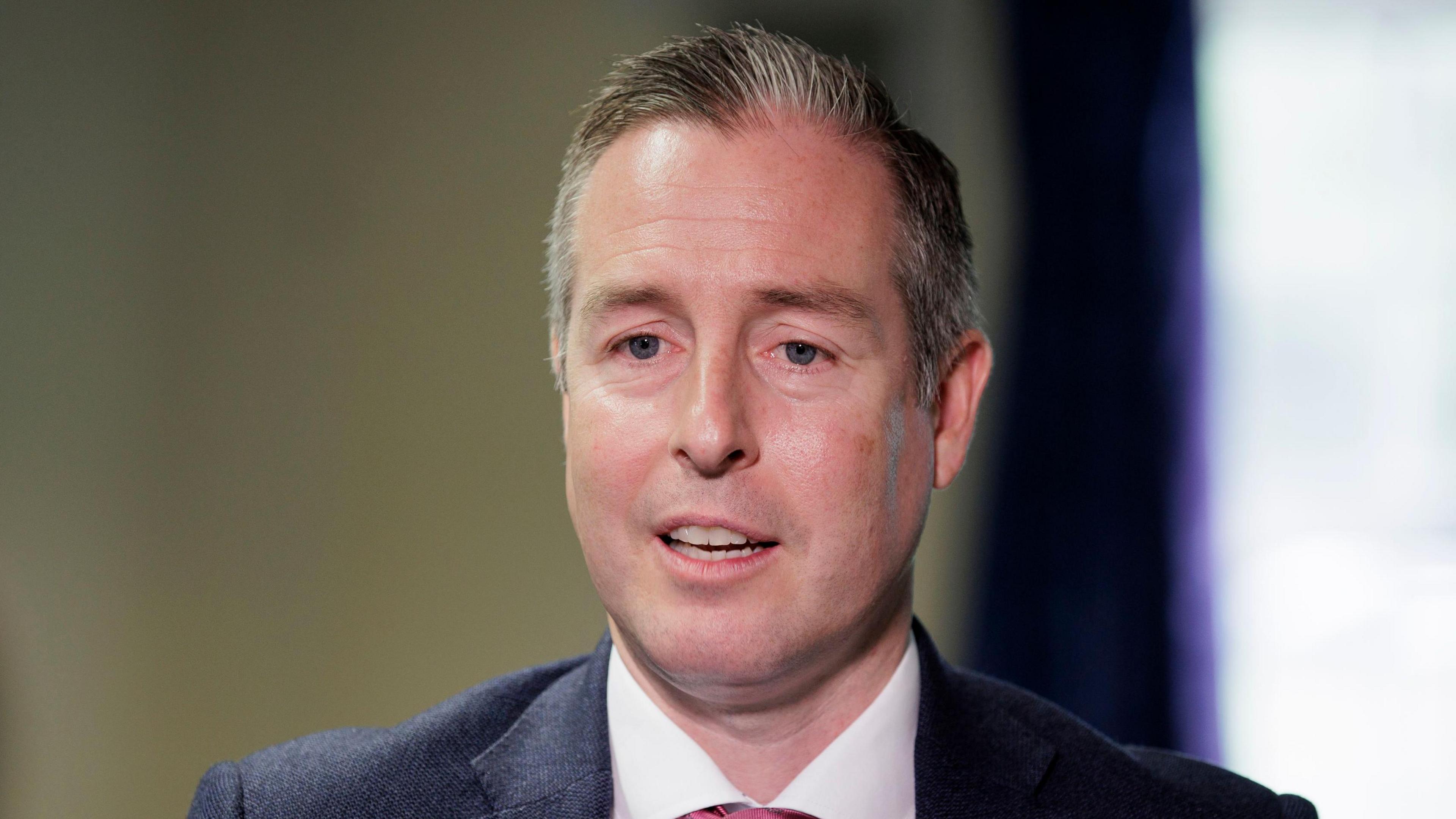 A man with short grey hair is talking to someone off camera. He is wearing a dark coloured suit jacket, a white shirt and a red tie. 