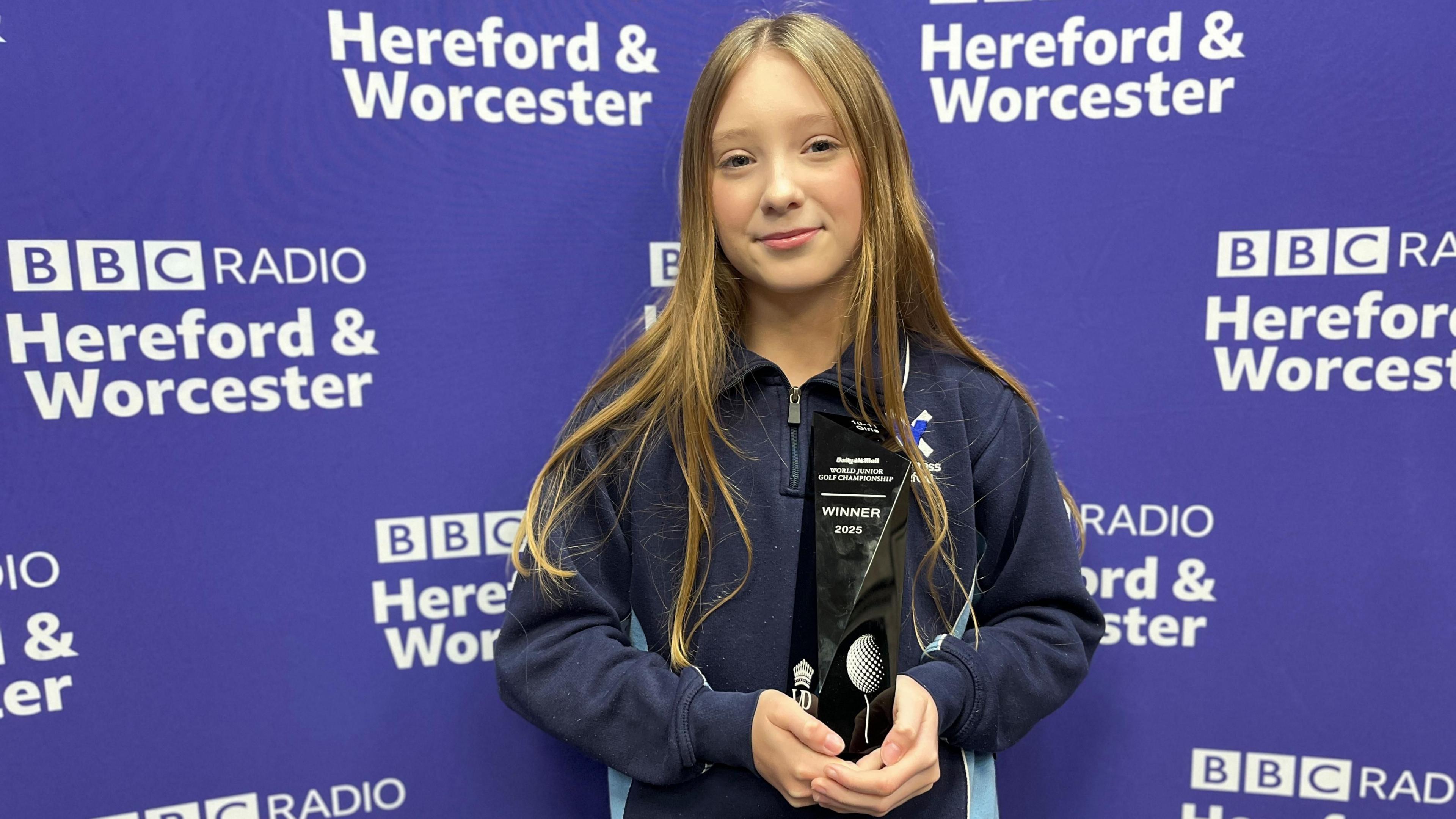 A young girl wearing a dark blue top stands in front of a purple banner decorated with the BBC Radio Hereford & Worcester logo. She is holding a black golf trophy which reads "Daily Mail World Golf Championship - Winner 2025."