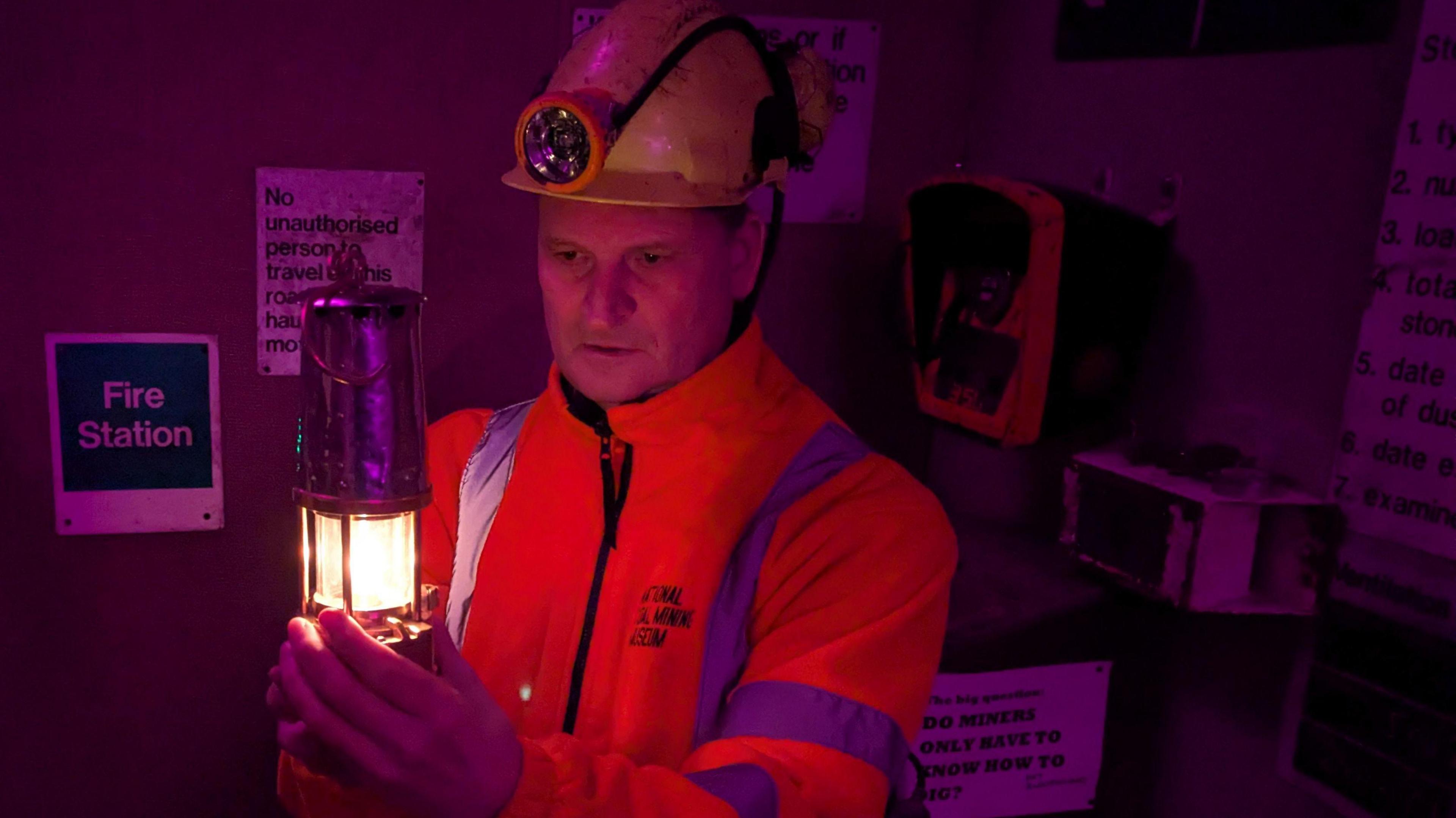 A man wearing a helmet and luminous orange jacket holds a mining lamp aloft. Various physical artefacts and signs are located on the shelves and walls behind him.