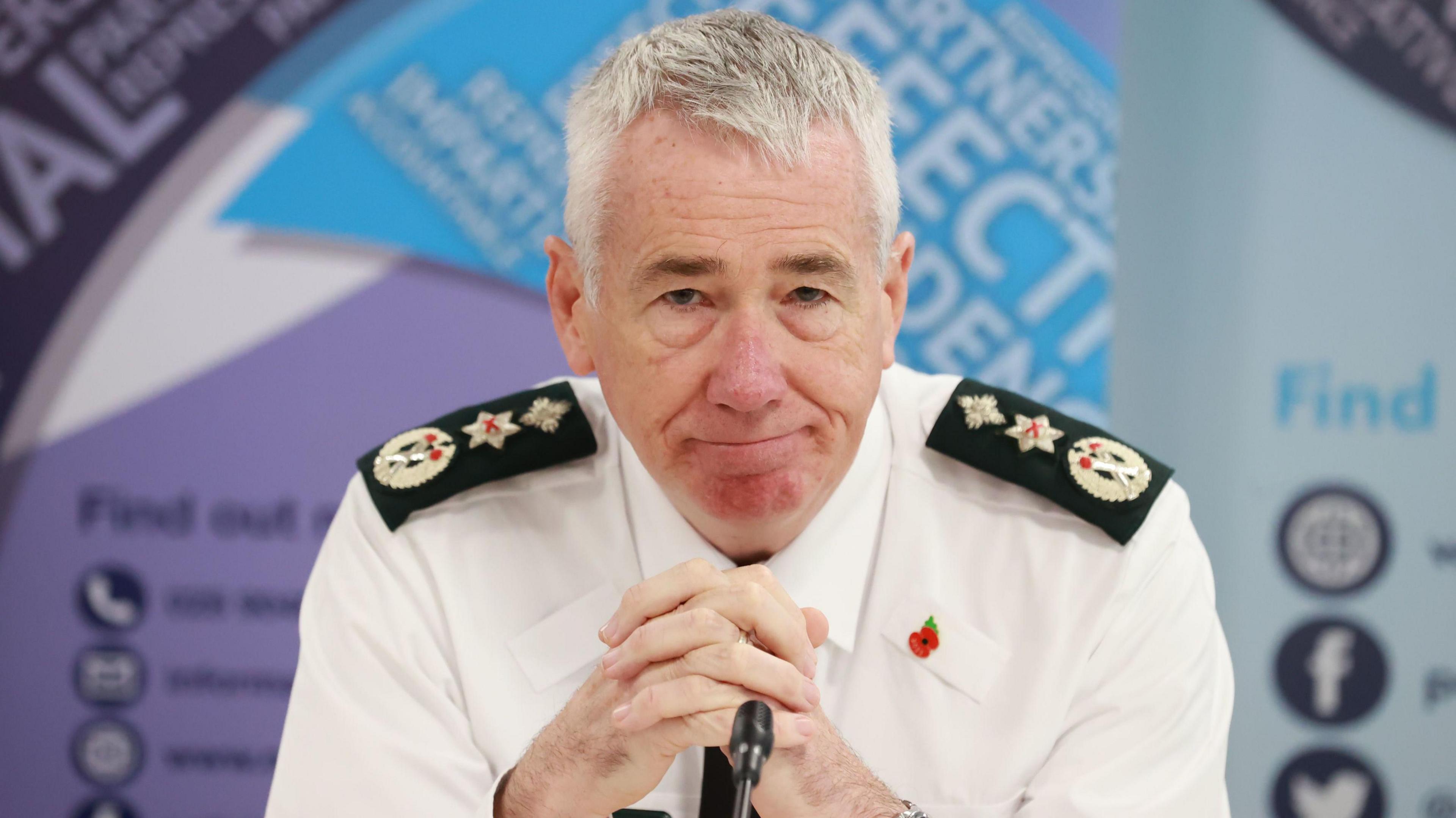 Jon Botucher sitting at a desk in front of a microphone with his hands clasped before him. He is wearing police uniform