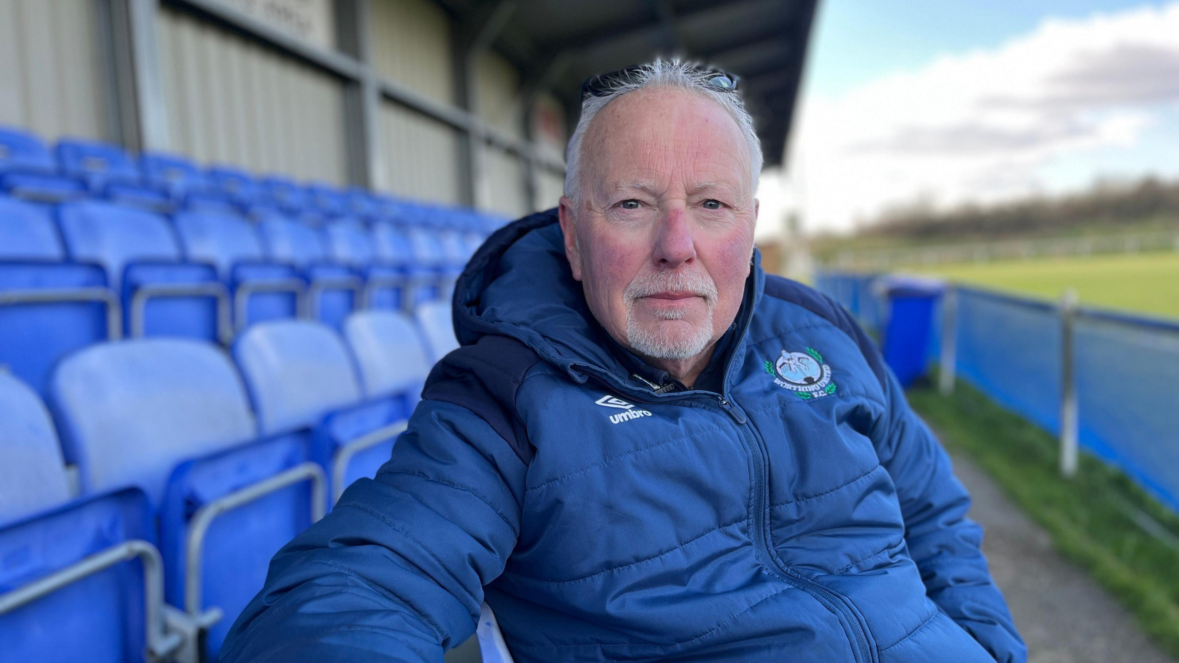 A man sits in a football stand. Behind him are blue seats. He is wearing a dark blue puffer jacket, has white hair and a goatee.