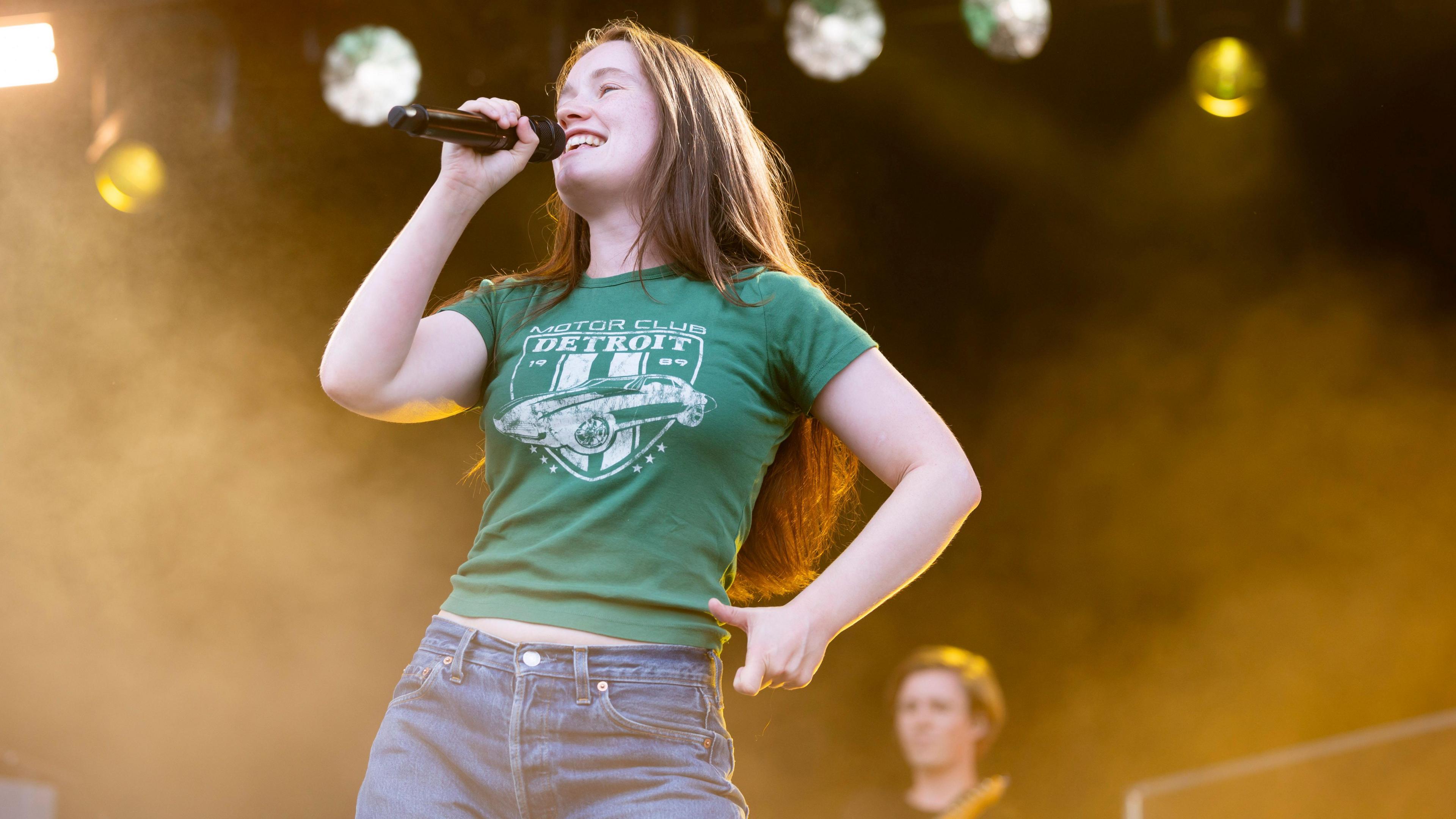 Sigrid on stage singing into a microphone. She is smiling, she has a short sleeved green t-shirt on, she has long light brown hair. Behind her above her head are stage lights