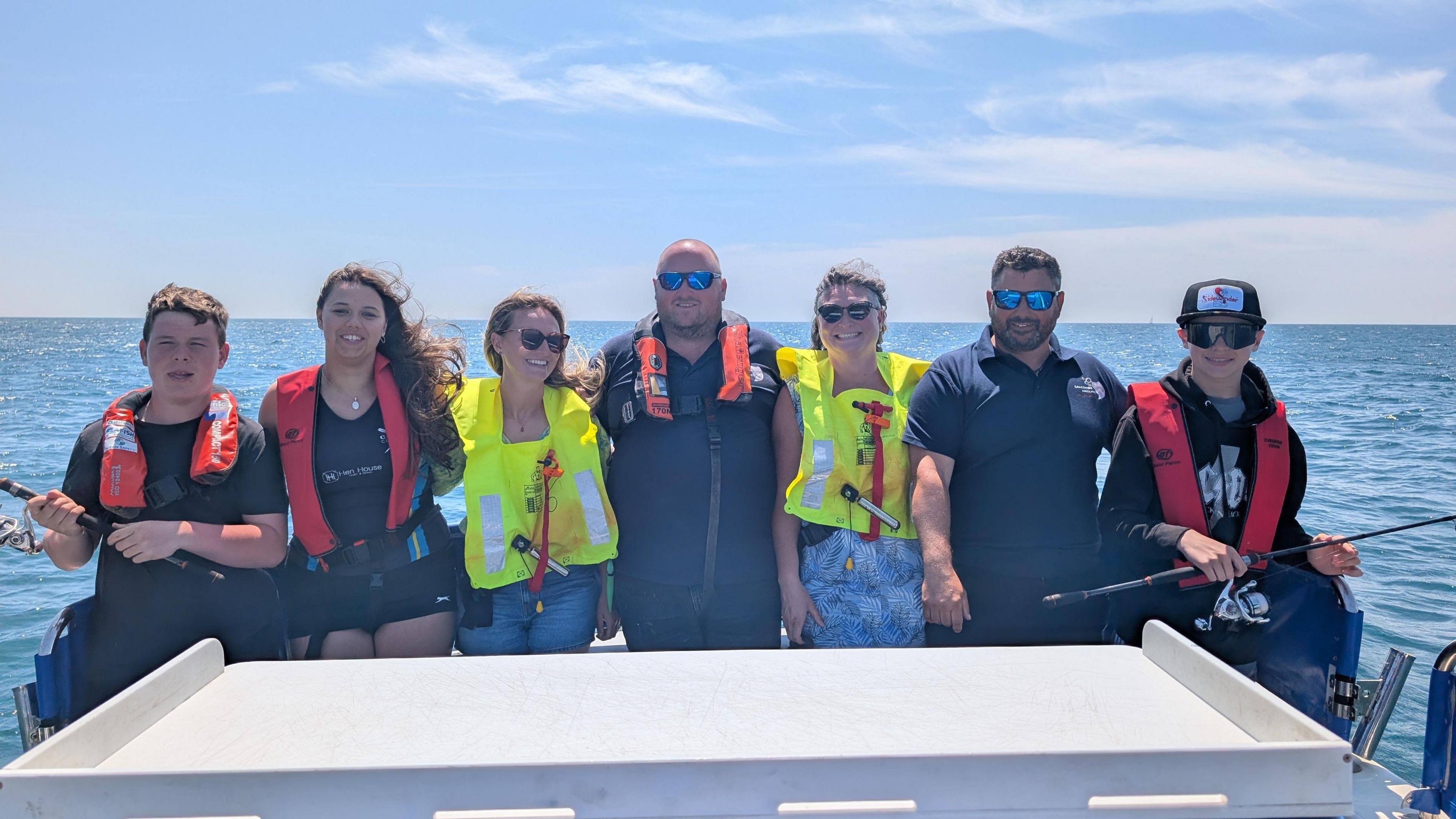 A group of people stand at the back of a boat in a row smiling at the camera with the sea in the background. They are all wearing lifejackets and the two teenage boys on either end are holding fishing rods.