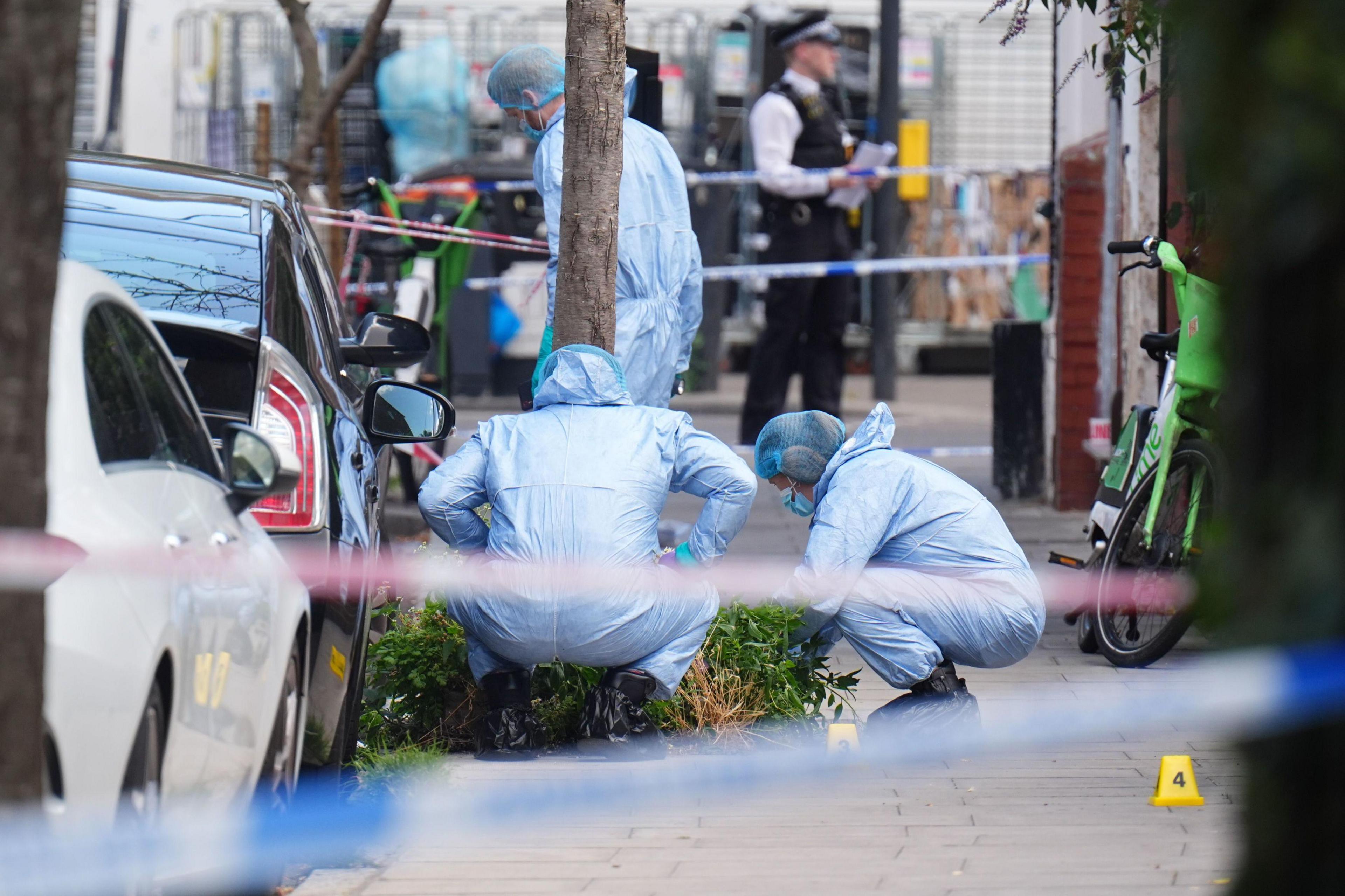 Three forensic police officers in blue overalls at the scene at Dynevor Road. Police tape, parked cars and a Lime bike can be seen