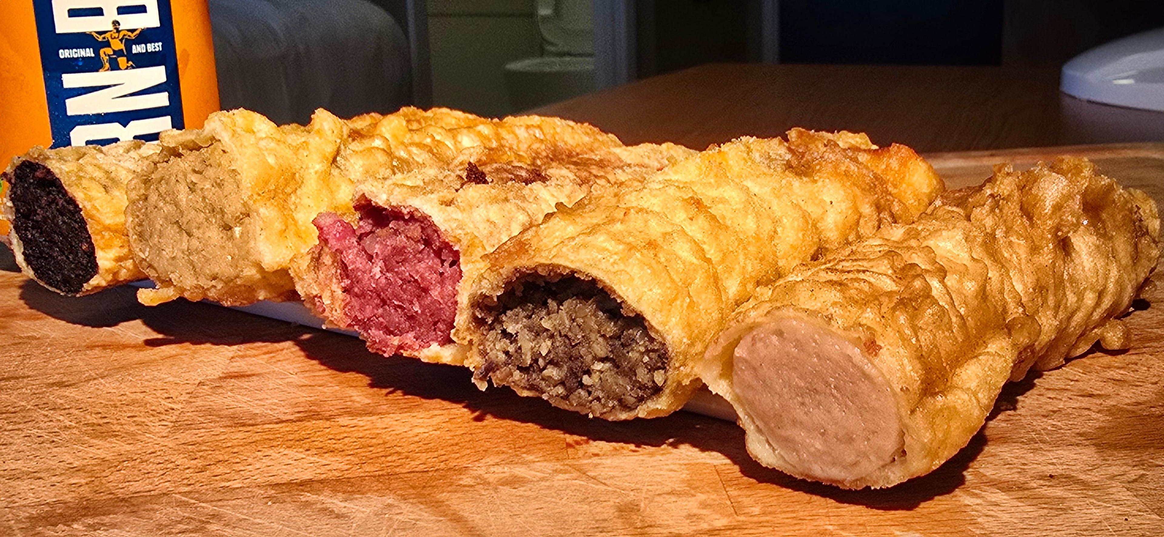 A row of delights on a kitchen counter, with the ends cut, exposing the insides of a black, white, red, haggis and a battered sausage. An orange Irn Bru can sits on the left.