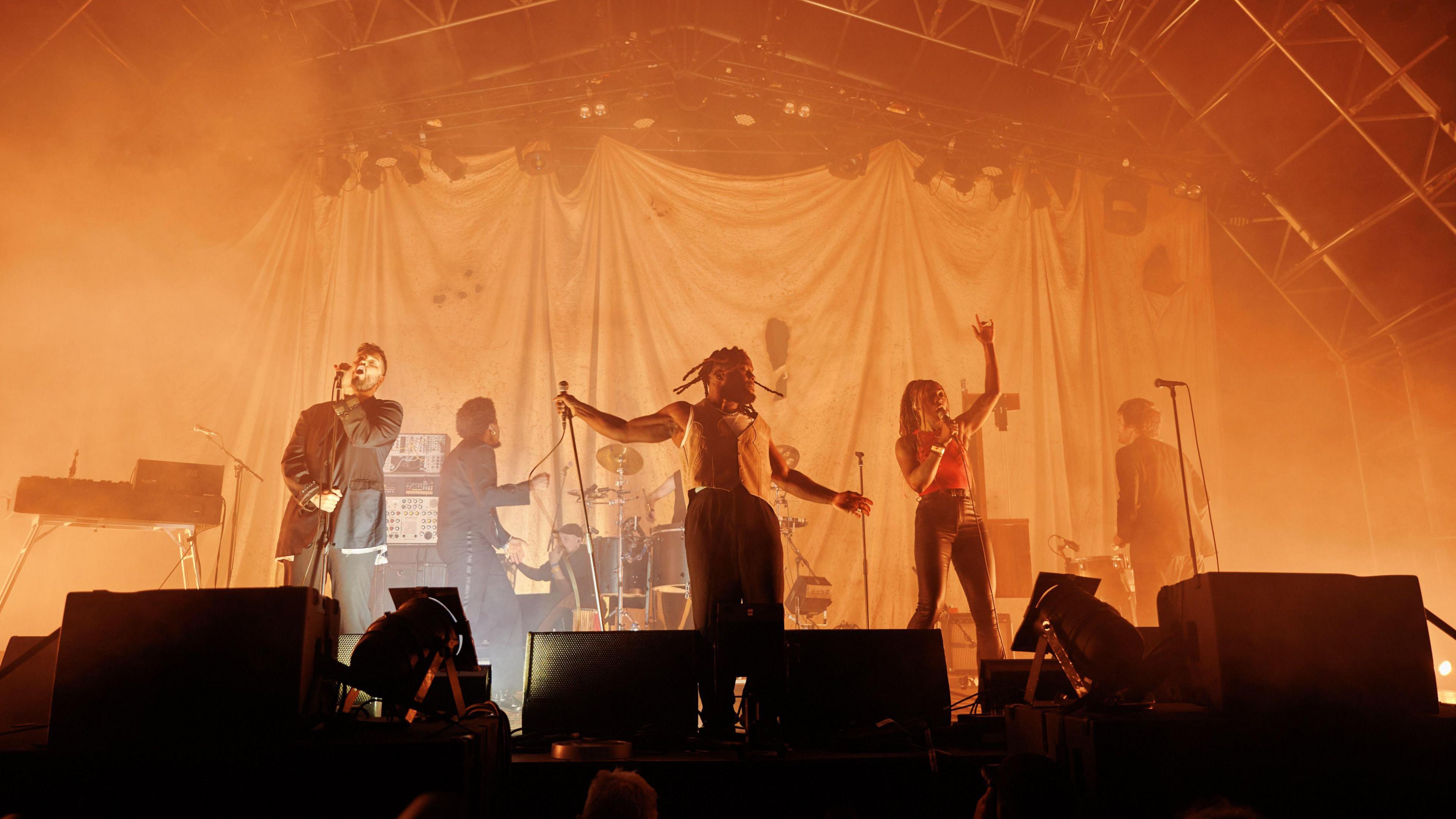 Six artists performing on a large stage in orange lighting