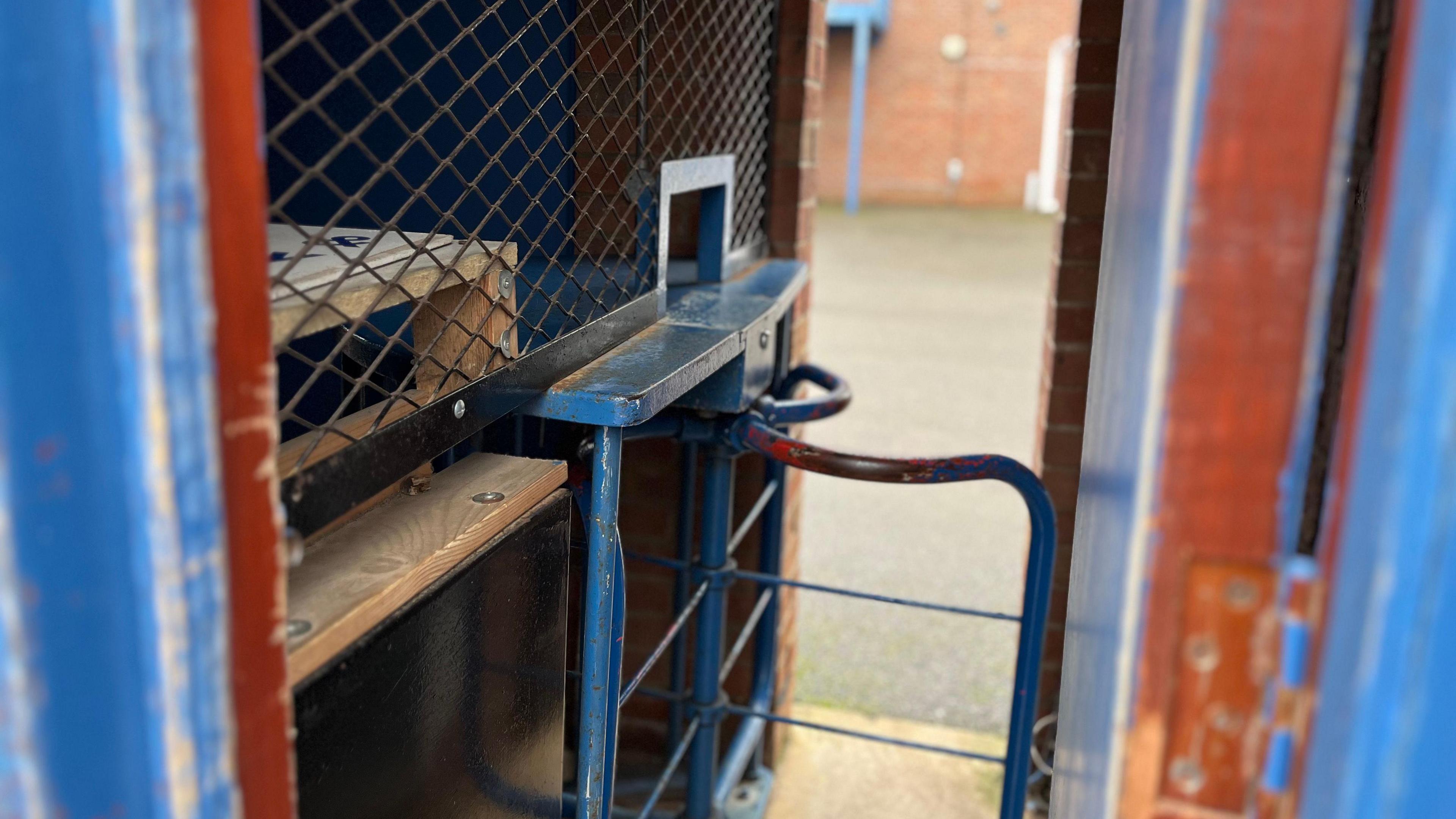 A very narrow outdoor turnstile, covered by a roof. Part of it has been painted blue, but parts of the paint have come away to reveal the dark wood underneath. The turnstile is a small blue gate. 