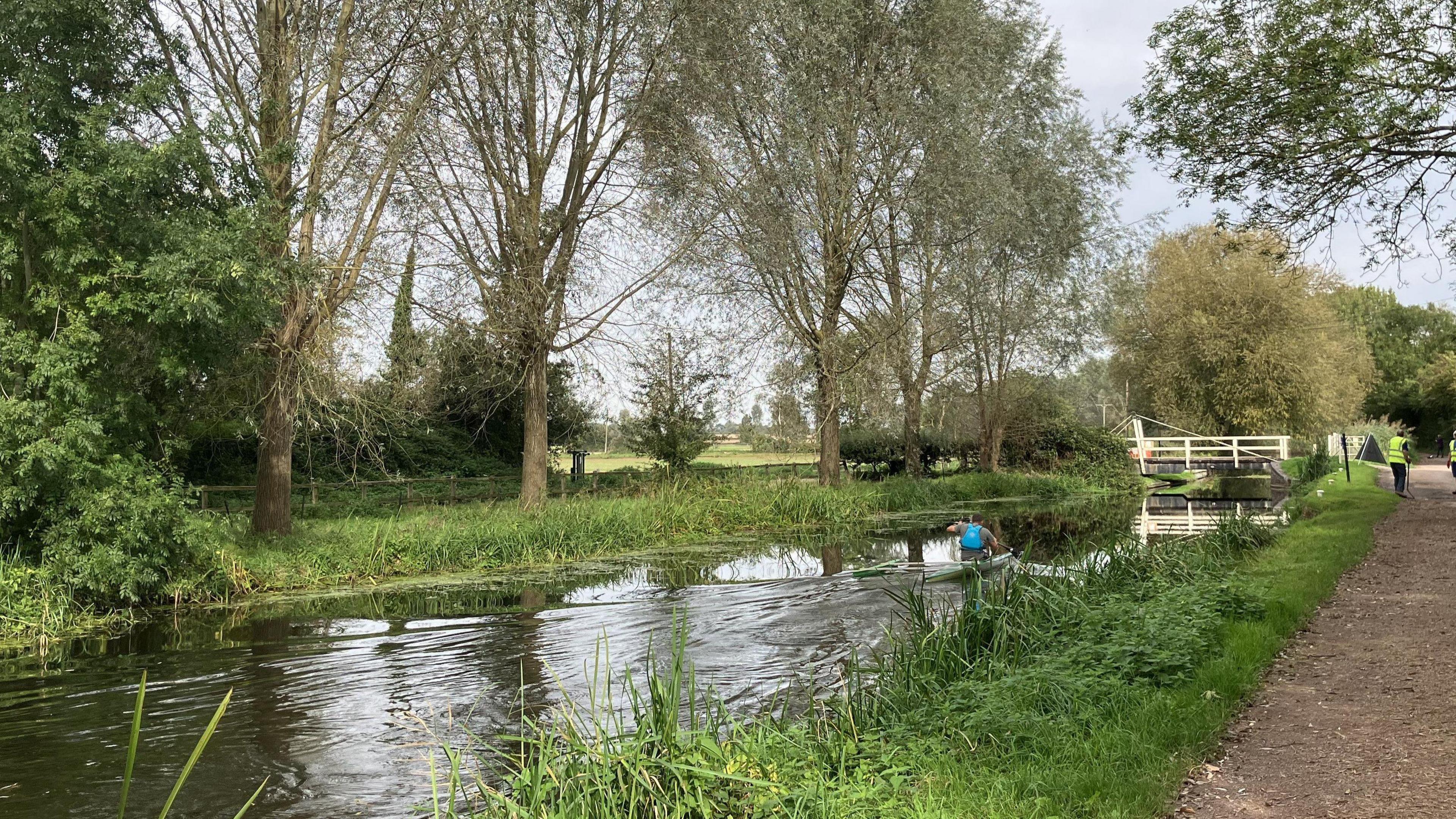 A kayaker on a canal with grassy banks and trees either side