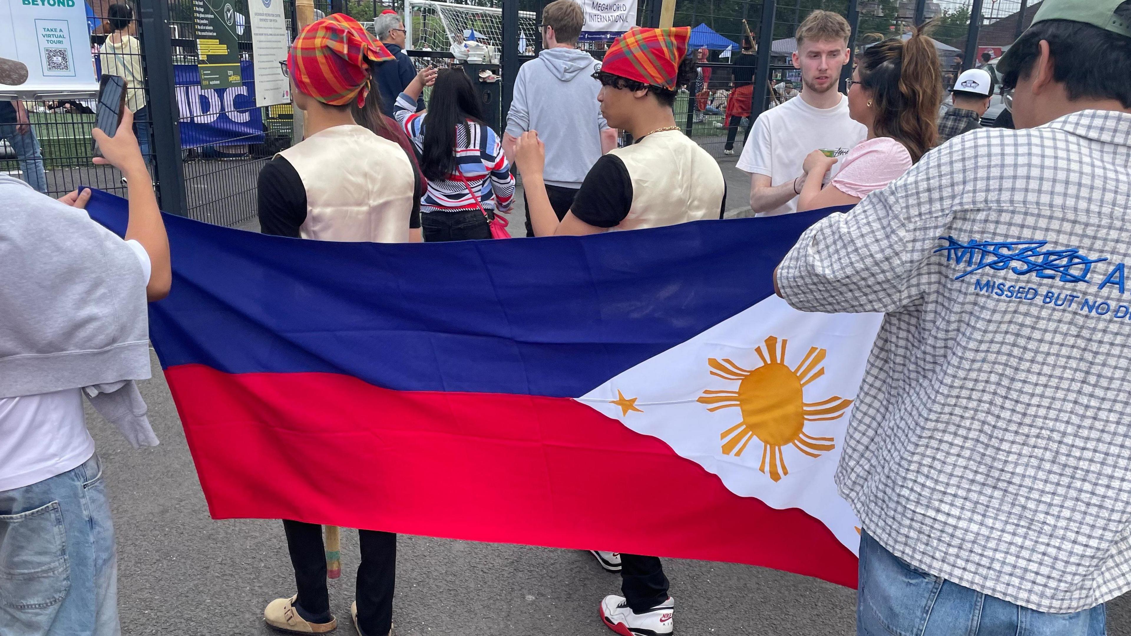 Two men hold a flag of the Philippines. It has a blue bar and a red bar underneath, a white triangle pierces the colours from the right with a yellow sun in the white part. Two men dressed in gold waistcoats and black t-shirts walk on the other side of the flag. The man holding the flag on the right wears a white and brown chequered shirt and green hat with blue jeans. The other man has on blue jeans and a white t-shirt, with a grey hoodie round his neck.