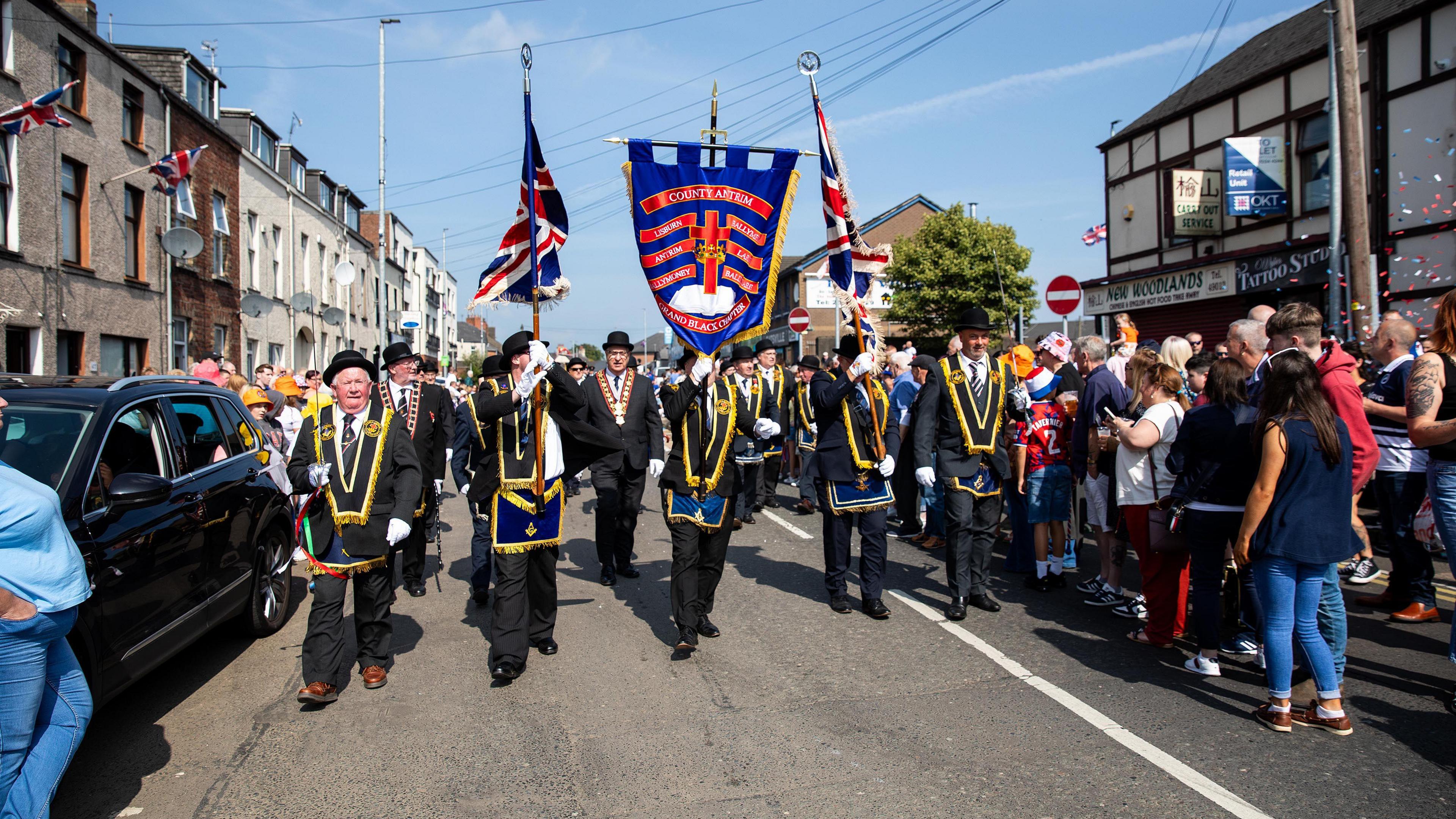 Members of the Royal Black Institution marching through a street in Ballymena, with members of the public standing on either side of the road.