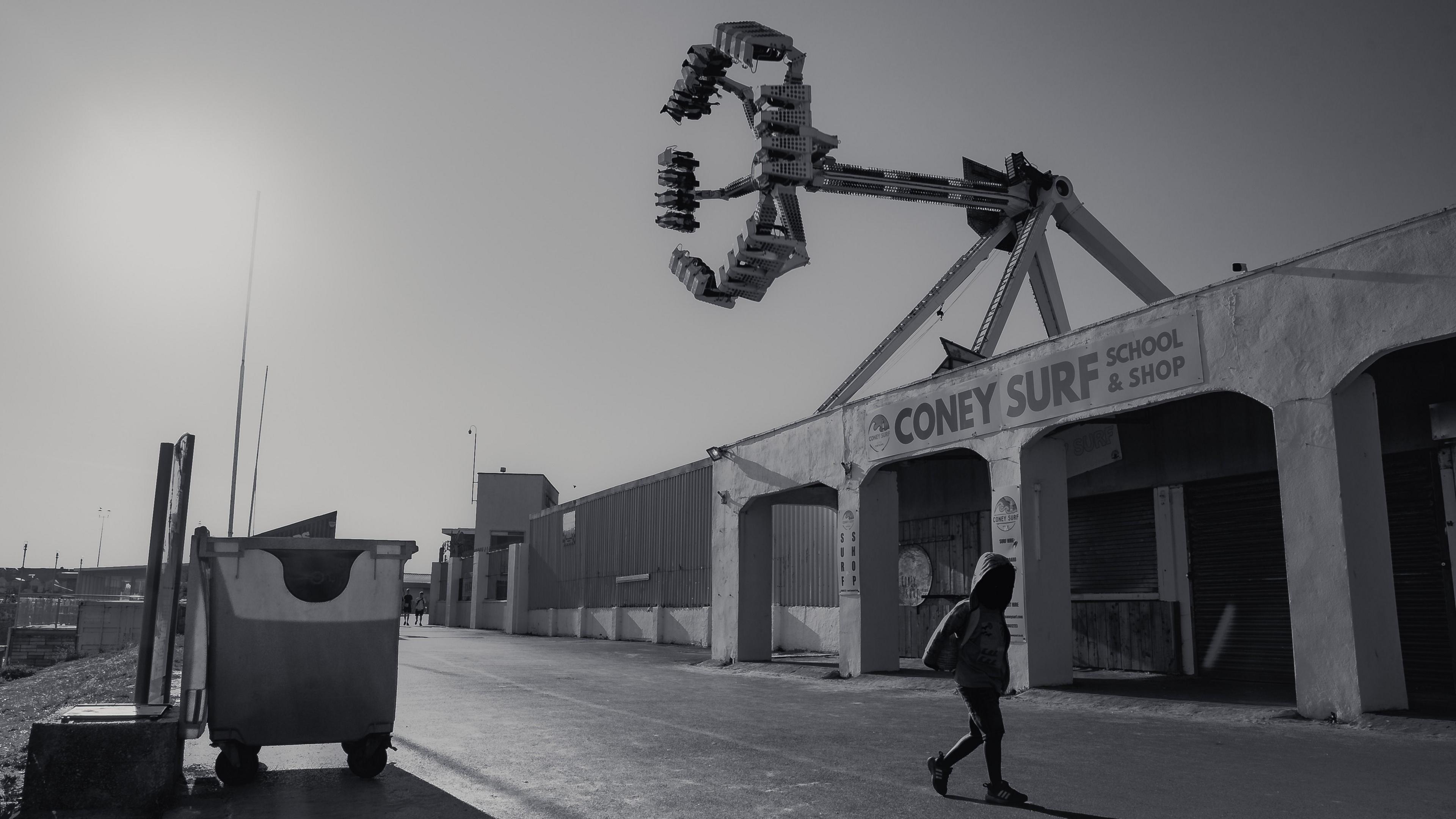 Black and white image of The 'Beach Party' ride has soared above Porthcawl promenade since 1999.
Black and white image of a revolving gondola fairground ride above a bleak concrete promenade, with the silhouetted figure of a young boy walking in the foreground. Coney Beach, Porthcawl, 21 August, 2025.