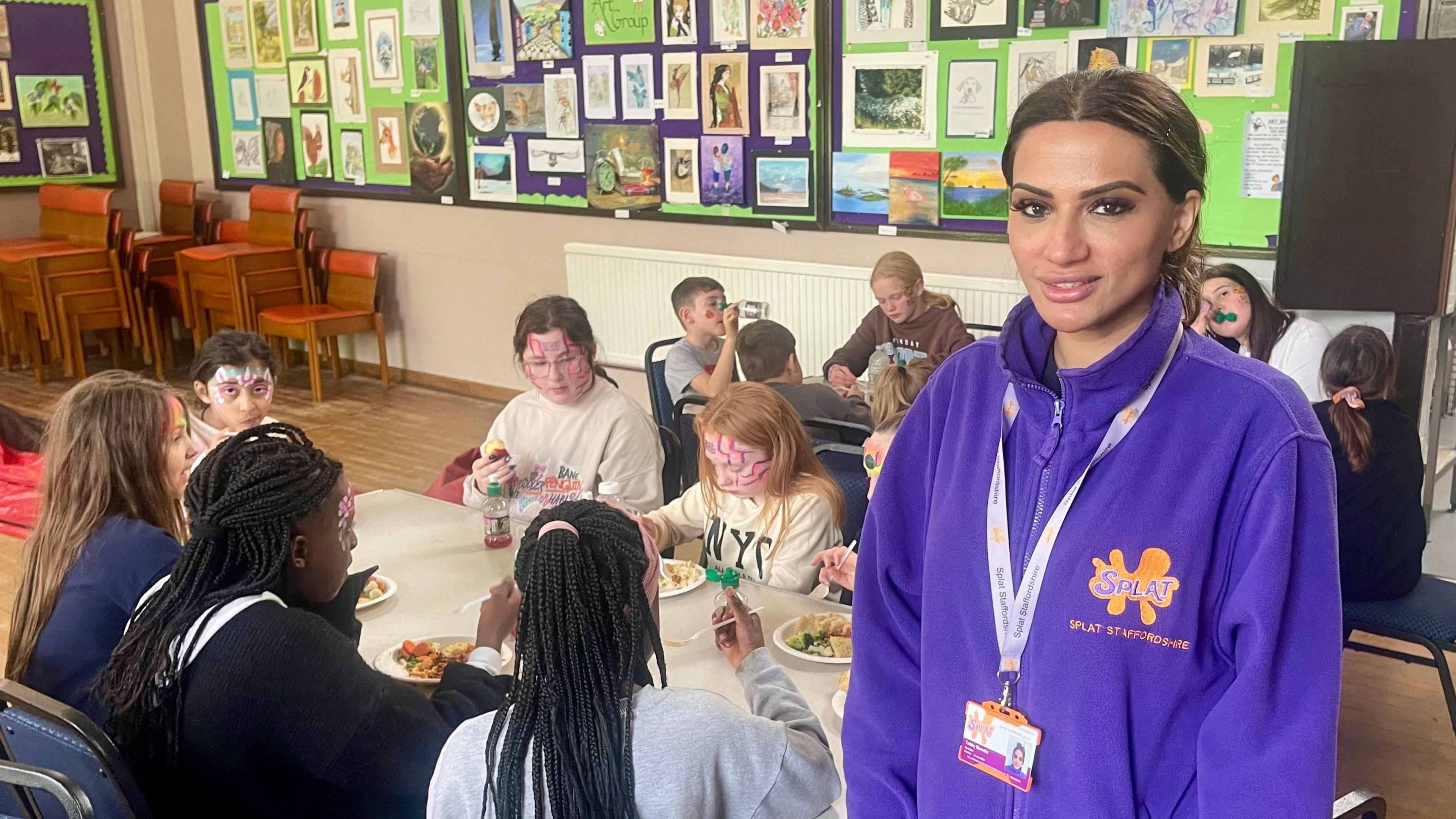 A woman in a purple fleece stands in front of two tables full of kids sat down eating spaghetti bolognase, garlic bread and vegetables. The woman has dark hair tied back, a white lanyard, ID badge and an orgnge logo embroidered on her left breast. There are wooden chairs stacked in the corner of the hall, with display boards covered in paintings and drawings on the wall.