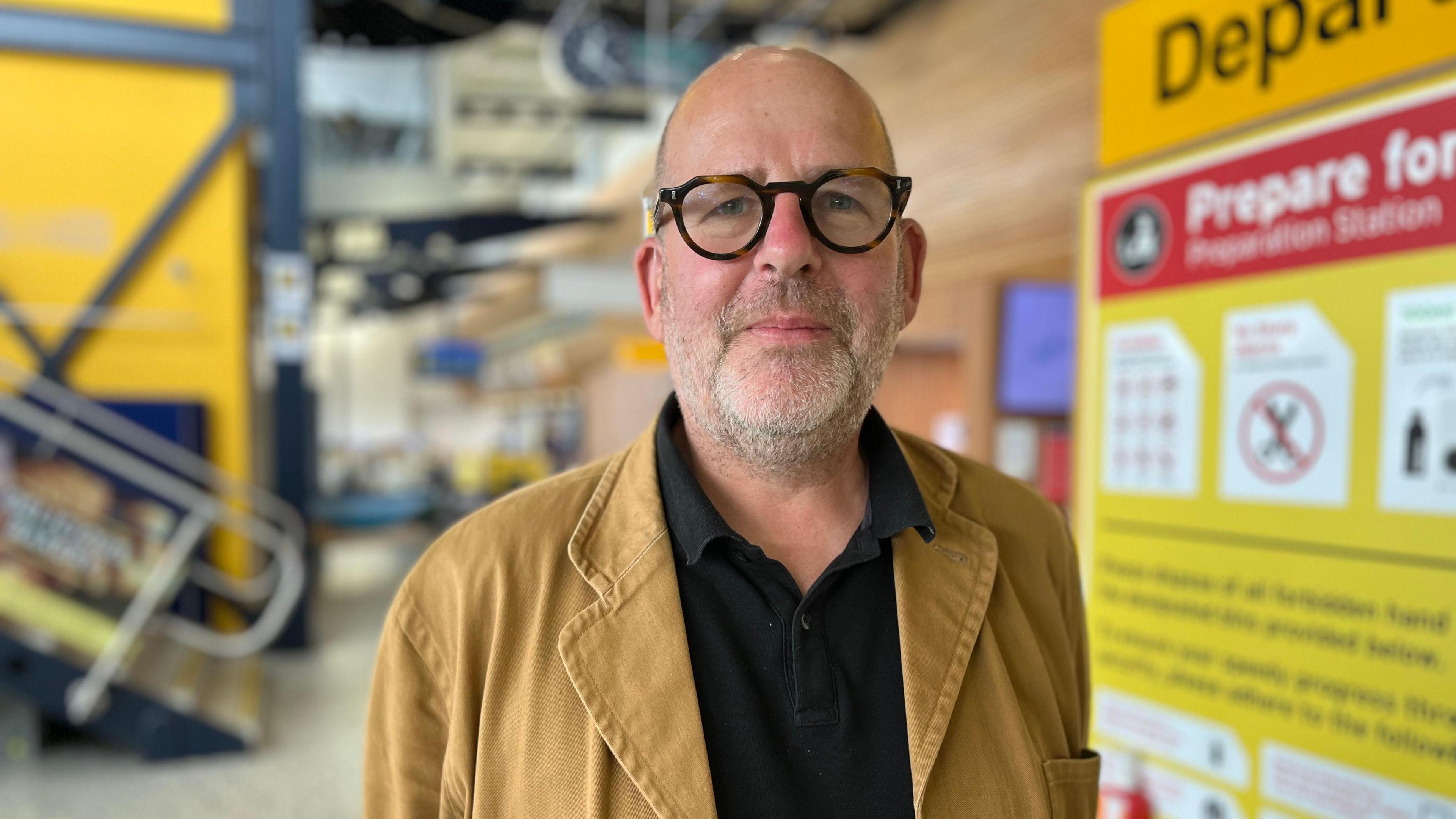 Professor Andrew Le Sueur is standing outside the security entrance next to departures. He has round tortoiseshell coloured glasses on. He is wearing a black t-shirt with a camel coloured jacket over the top