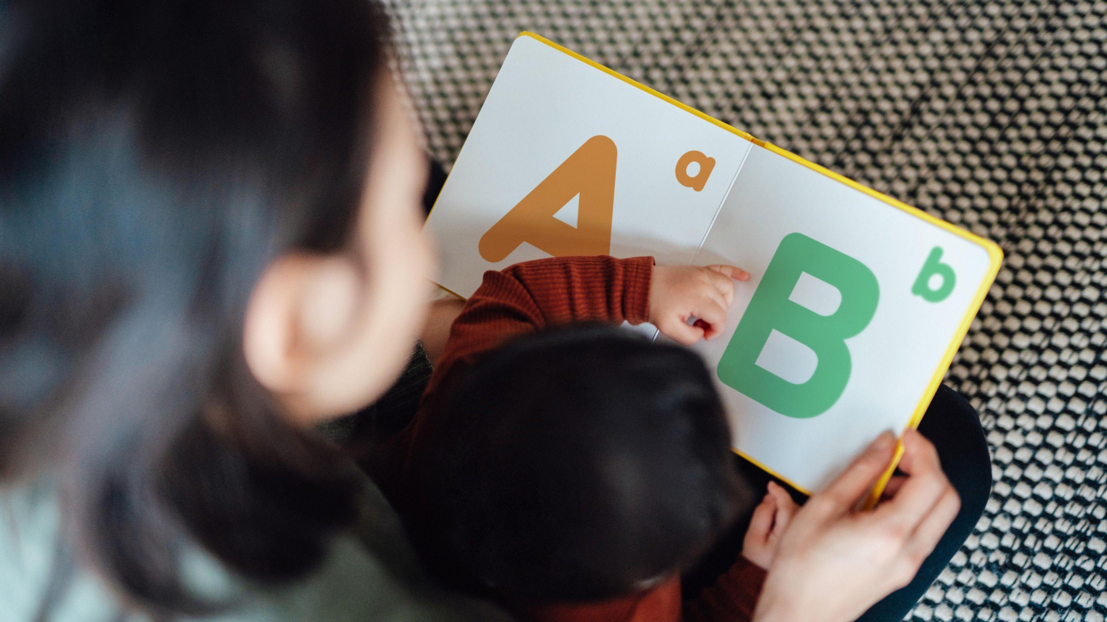 A blurred image of a parent reading an early learning alphabet book to her young child.