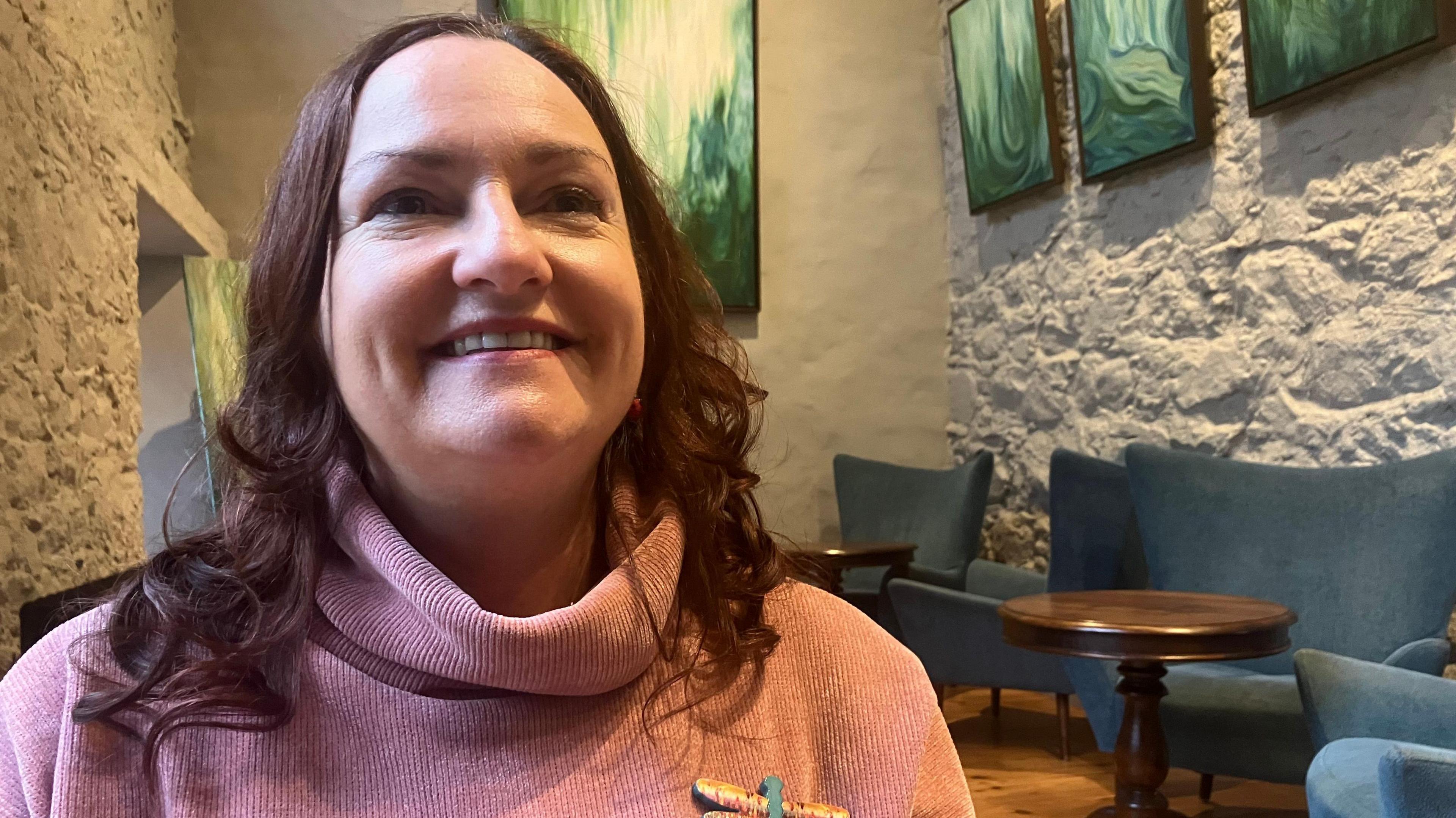 Ann Ward is smiling and looking away from the camera. She has curly hair and is wearing a pink turtleneck jumper and a dragonfly brooch. She's standing in a café. 