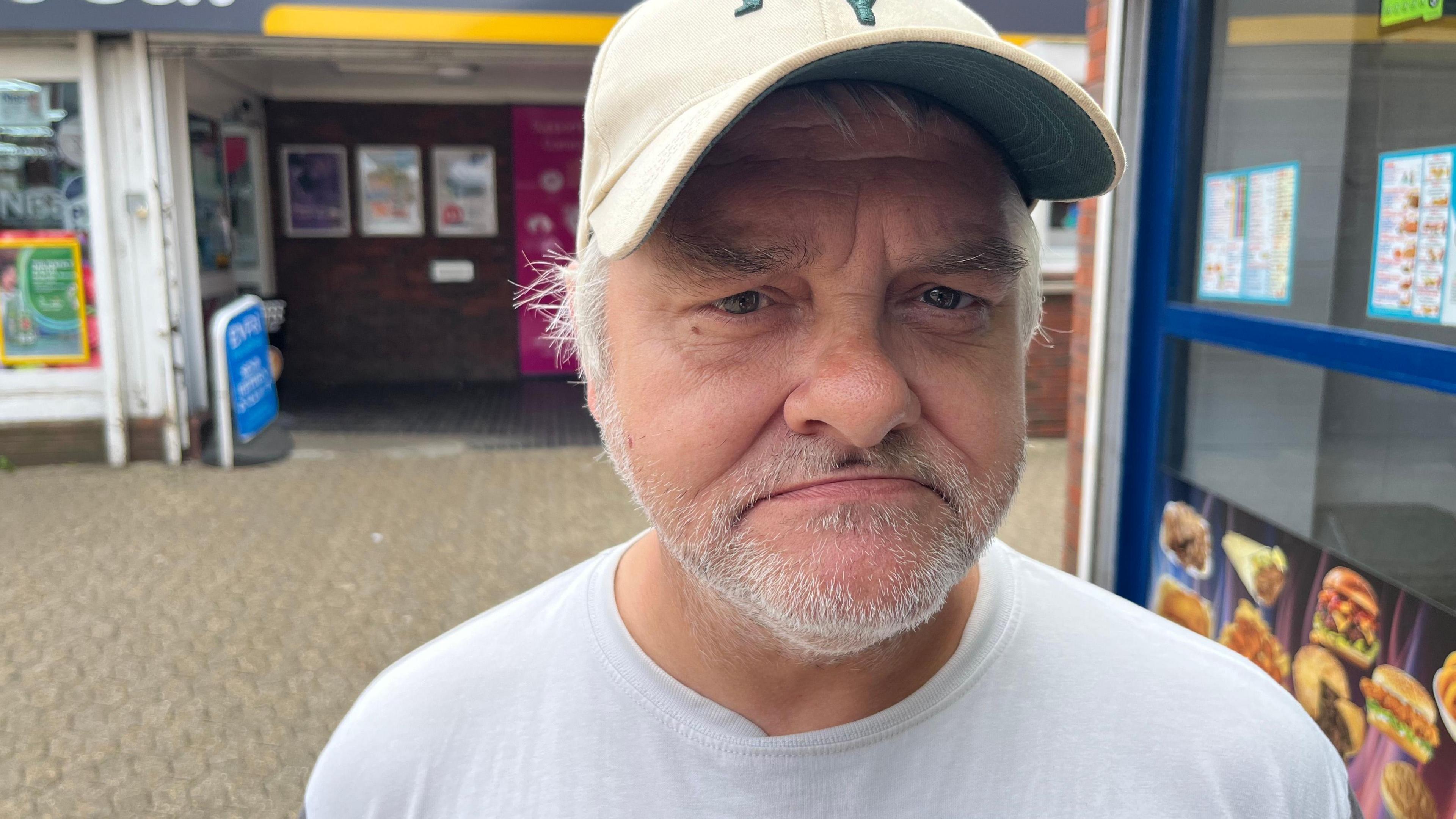 Tim Clark is frowning and looking towards the camera. He is wearing a white cap and white t-shirt. Behind him is a newsagents and fast food restaurant.