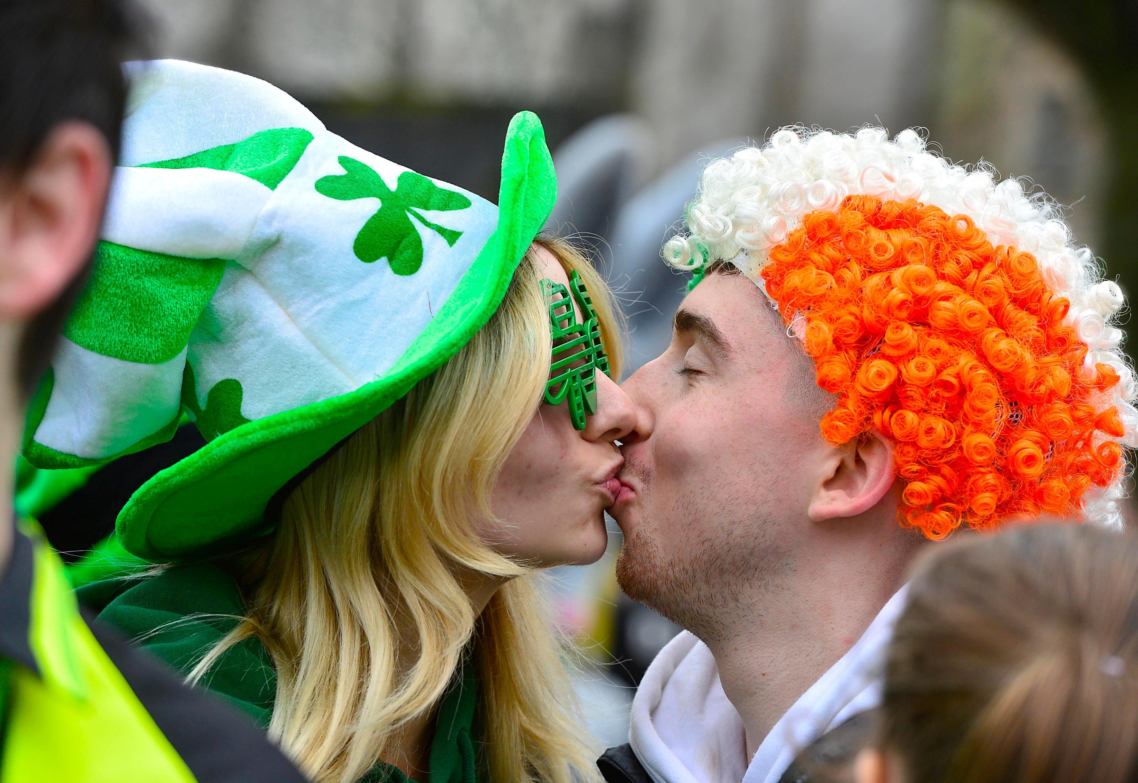 a woman wearing shamrock glasses and a St Patrick's Day hat kisses a man in a green, white and orange wig