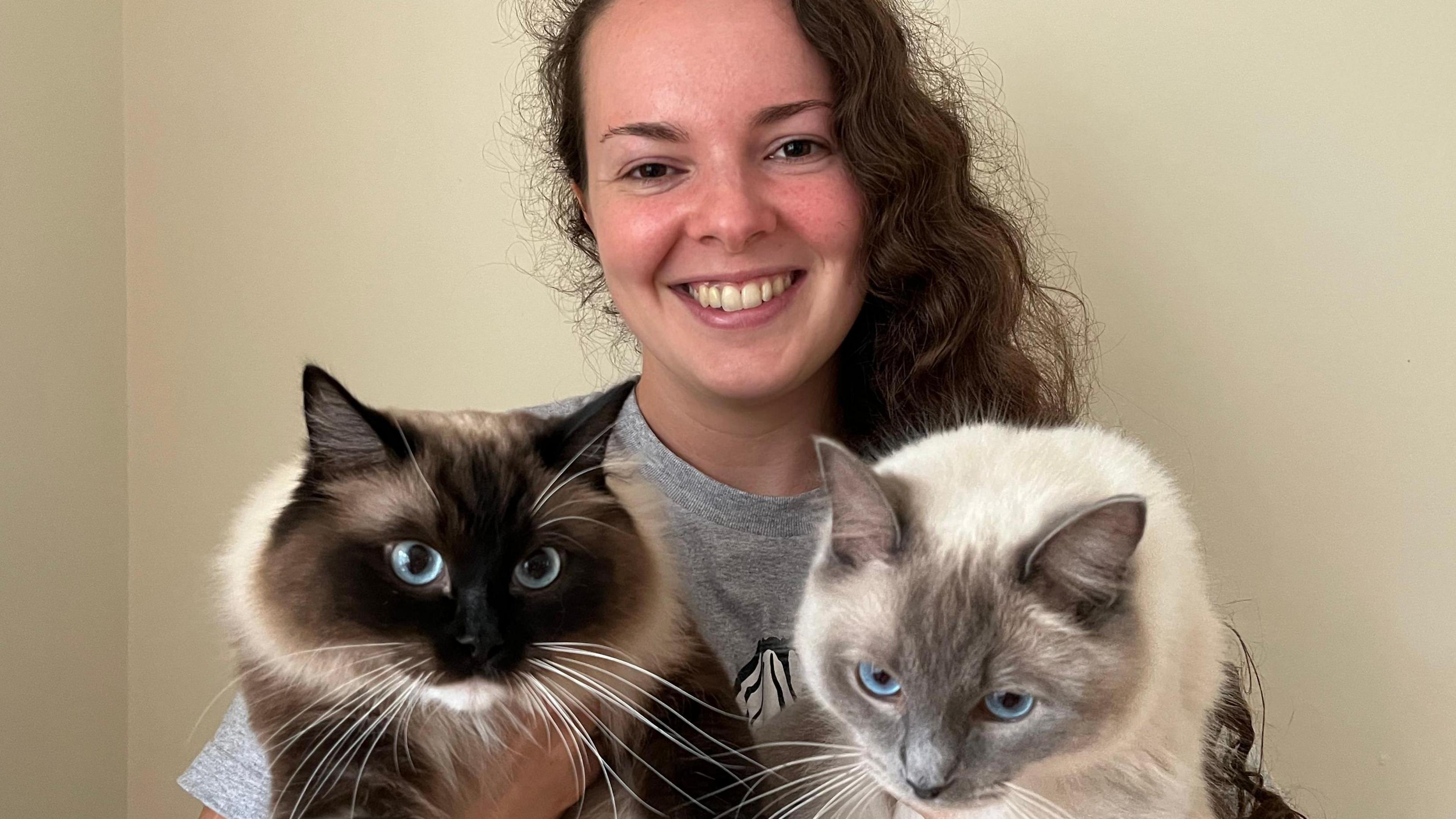 A picture of a lady wearing a grey shirt with black hair, holding two cats. The cat on the right of the image is black and cream in colour. The cat on the right is grey and white.