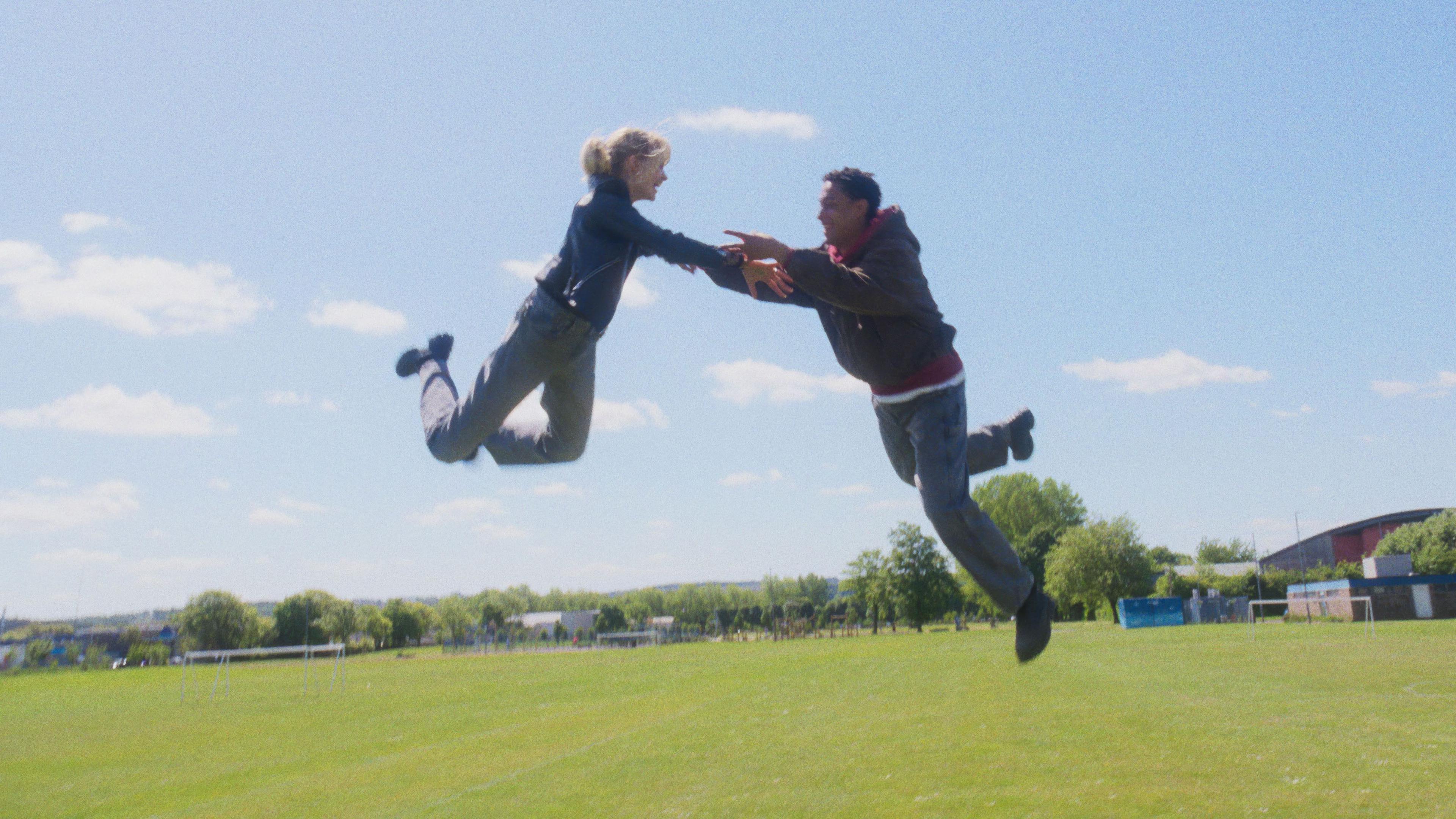 A woman wearing a blue jacket and grey trousers floats in the air, she's holding hands with a man wearing jeans and a red and grey jumper who is also floating