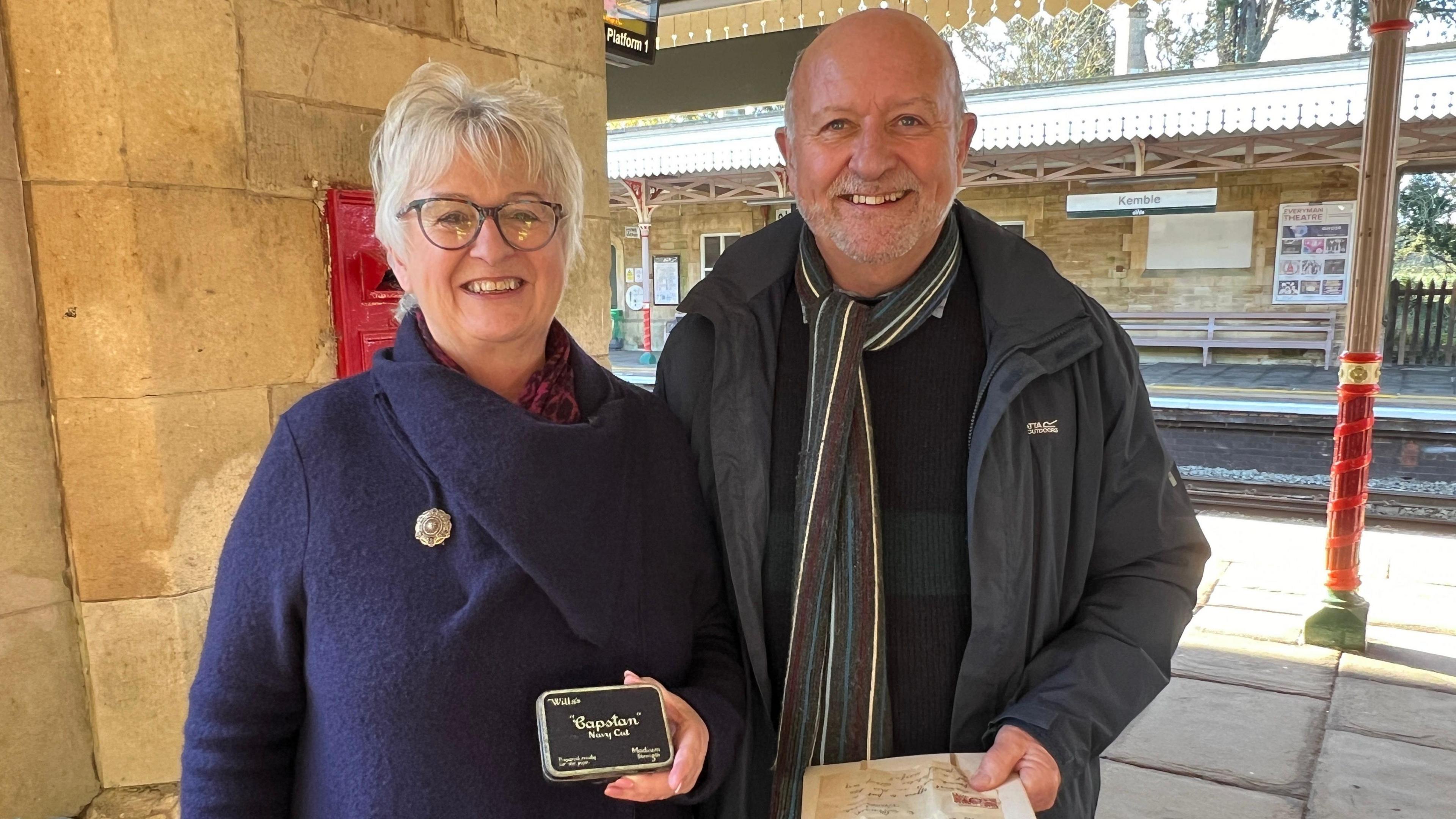 A brother and sister, probably in their 60s, stand on a railway platform on a cold, sunny day. The woman, who is wearing a blue woollen coat, holds an old cigarette tin, while the man holds an old letter and is wearing a striped scarf and a dark grey coat. They are both smiling.