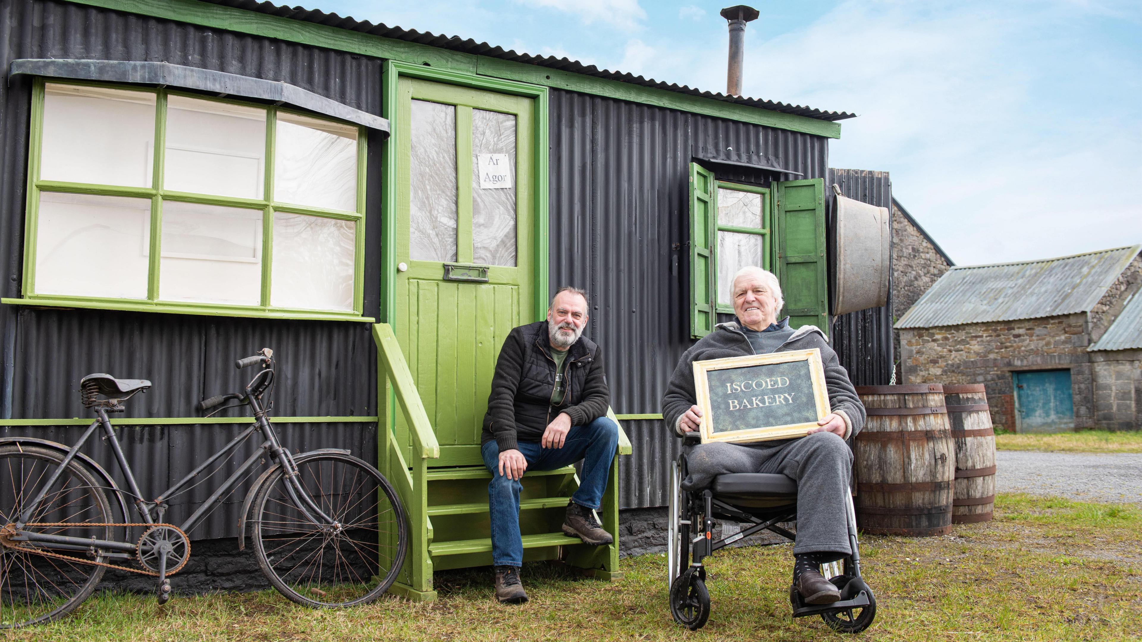 Seimon and his dad Stephen sat outside their mobile museum. Seimon is sat on the steps, while Stephen sits in his wheelchair to the right. He is holding a sign saying Iscoed Bakery. This side of the museum is a replica of Seimon's great-grandmother's bakery.