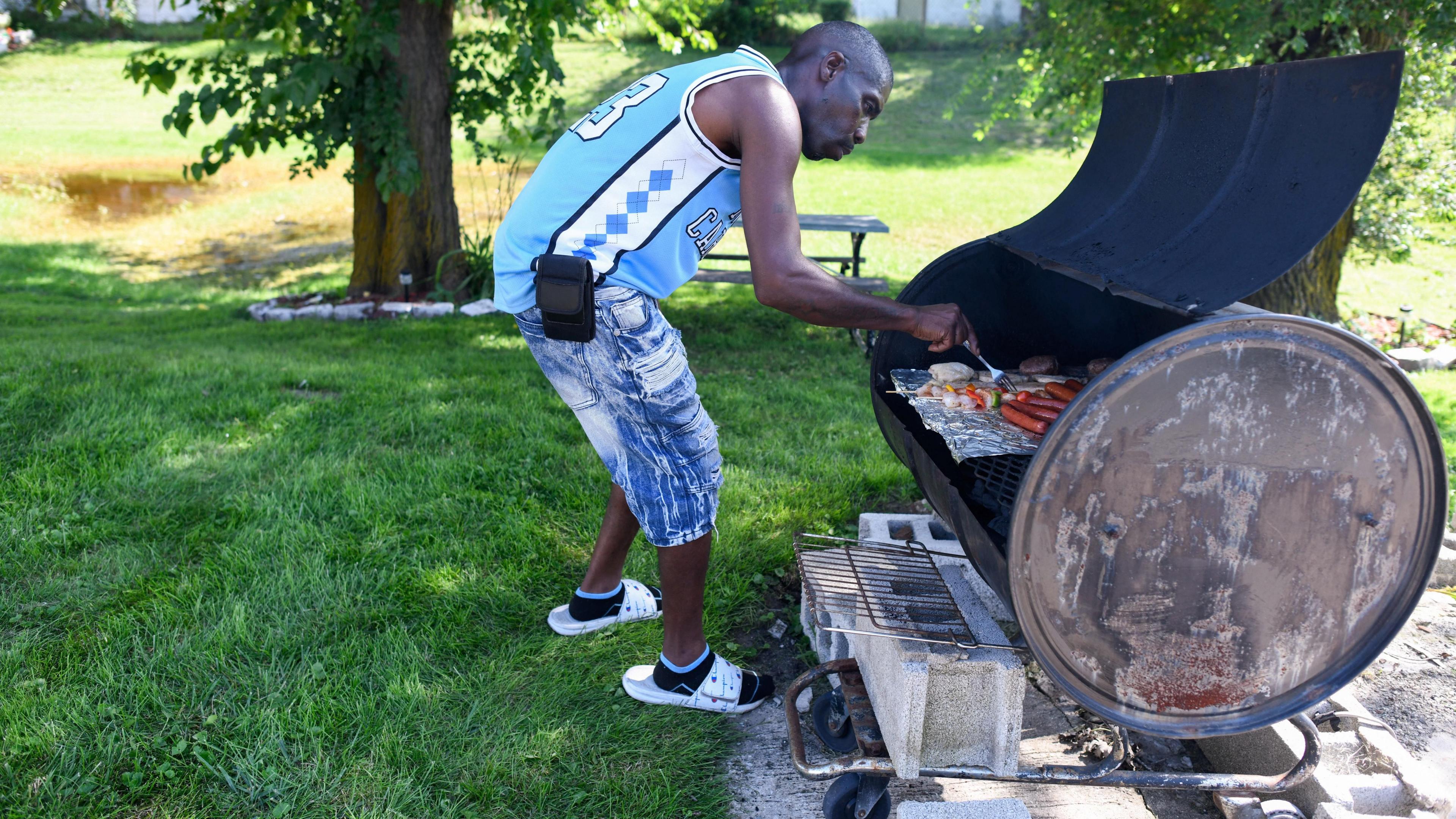 A man in a blue sports vest and jean shorts grills food in a garden