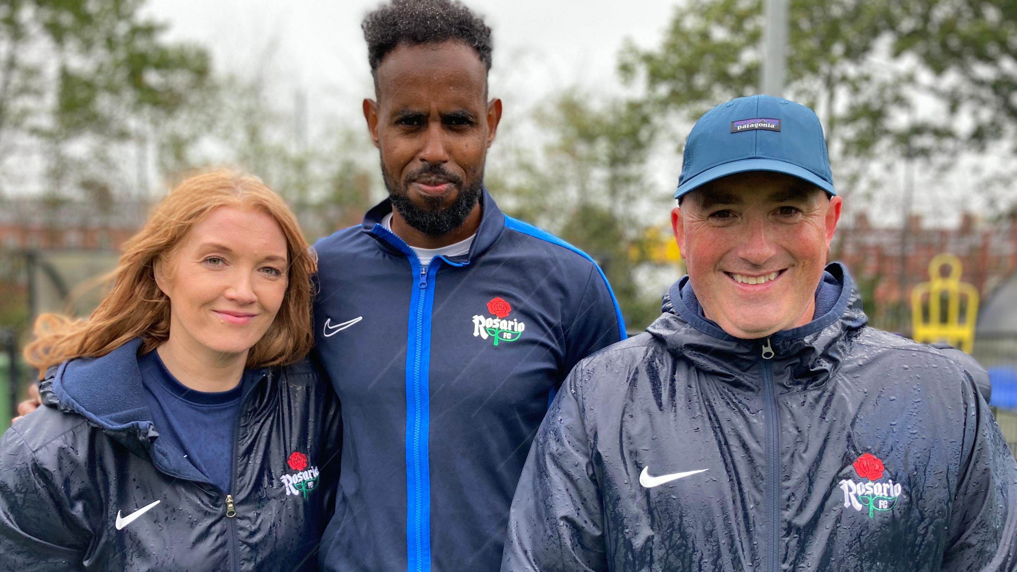 A lady with long red hair wearing a navy rain coast with a white nike tick and a rose logo with Rosario FC written on it. A man with black hair and a goatee beard wearing a navy rain jacket with a white Nike tick and a rose logo with Rosario FC written on it. A man with a light blue cap and a navy rain coast with a white nike tick and a rose logo with Rosario FC.