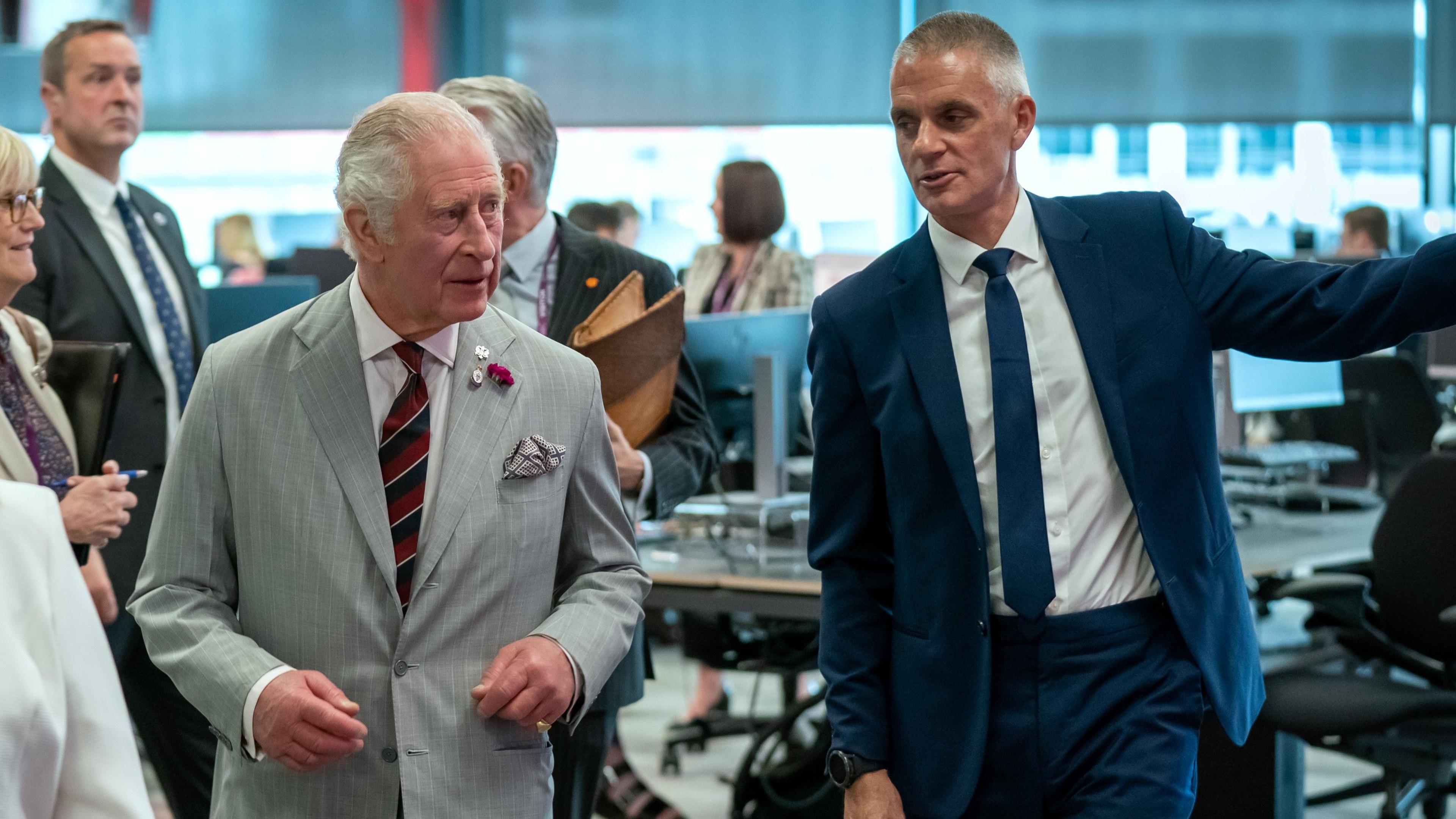Tim Davie gestures as he walks alongside King Charles through an office 